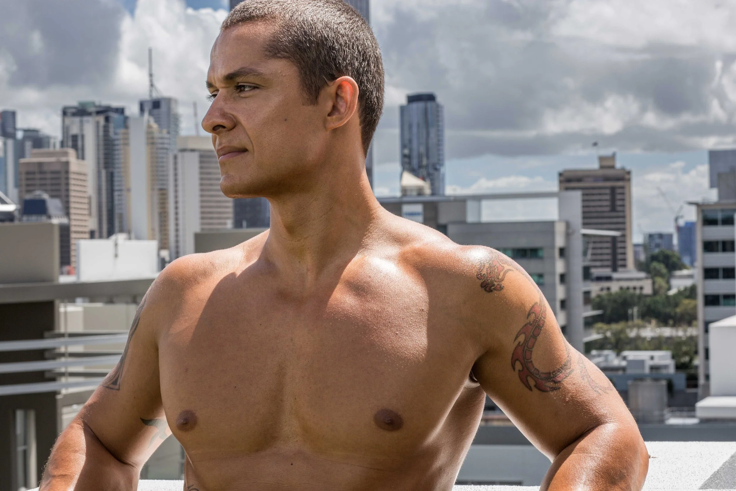 A shirtless man standing on a rooftop with a city skyline and clouds behind him.