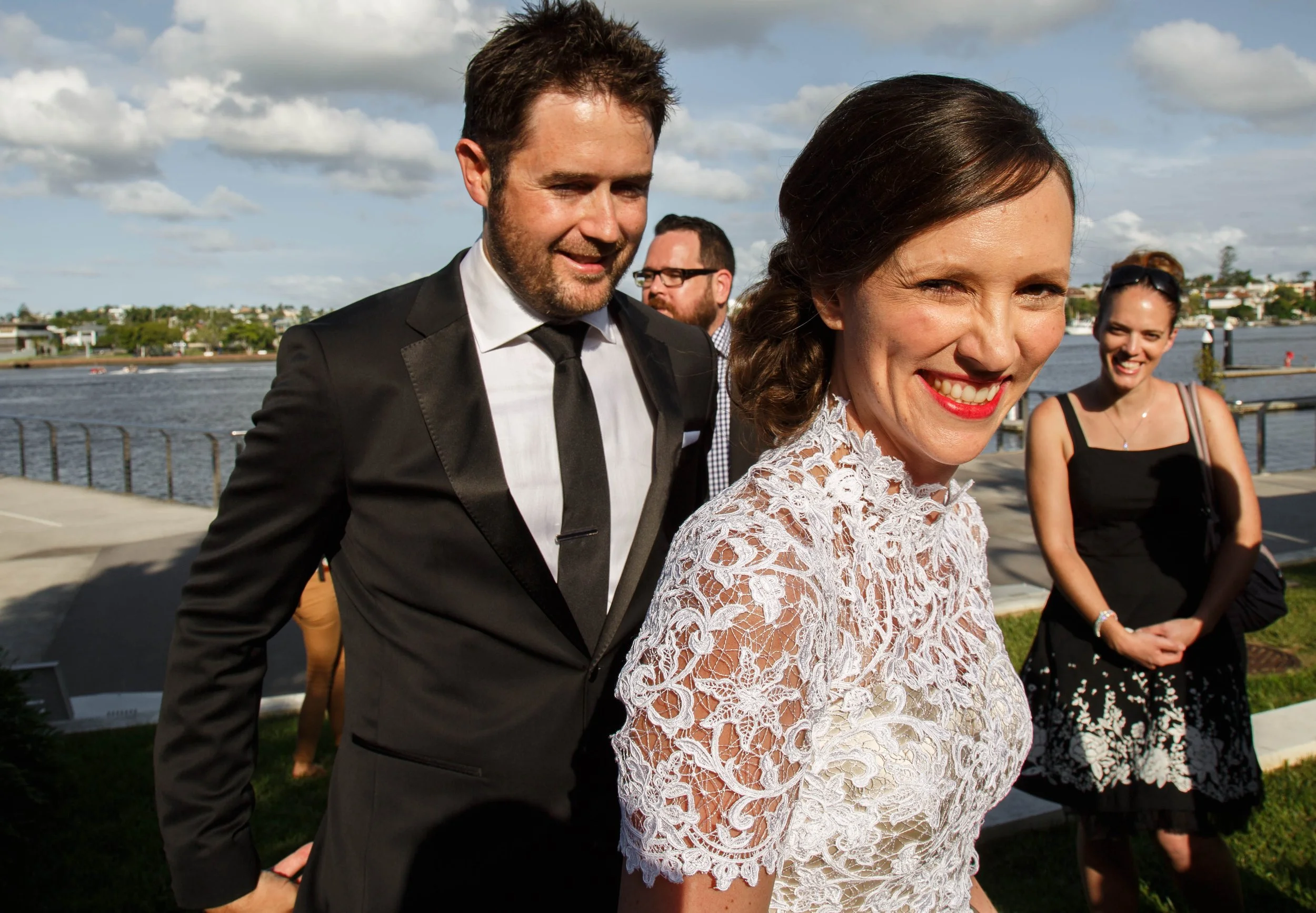 People at a waterfront outdoor event, including a smiling woman in a white lace dress, a man in a black suit, and a woman in a black dress in the background.