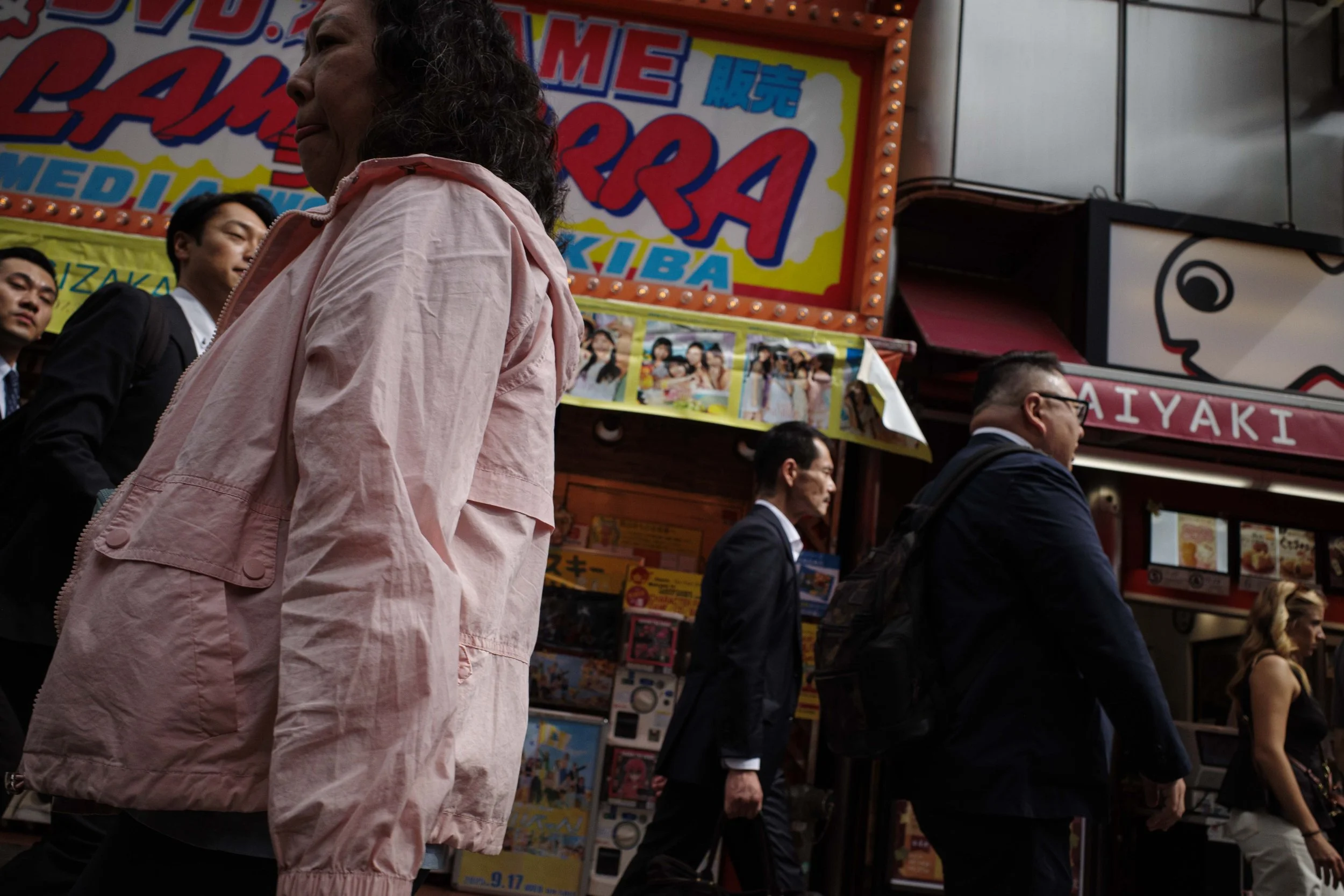 A crowd of people walking past colorful signs and shops on a city street, including a pink jacket woman in the foreground, and businesspeople in suits.