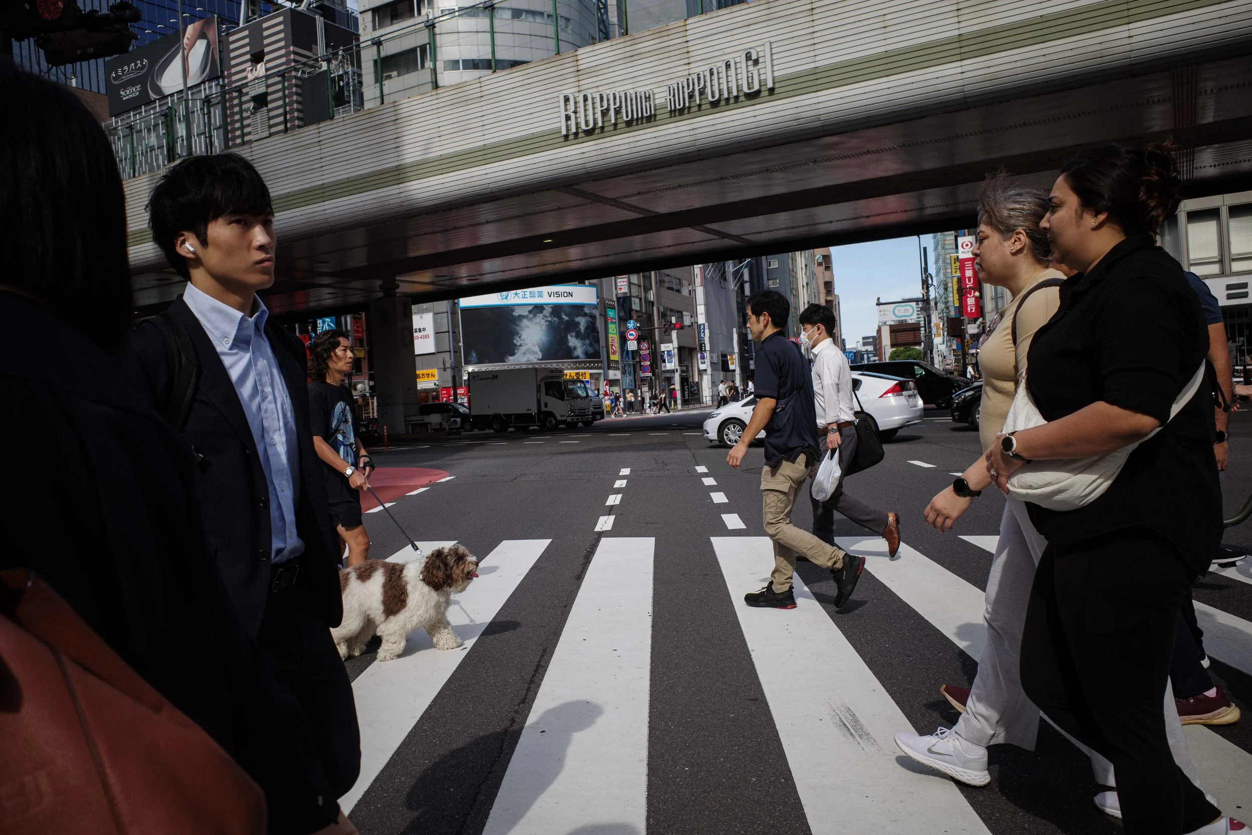 Pedestrians crossing the street in Tokyo under an elevated train station with the sign Roppongi Roppongi, surrounded by buildings and vehicles.