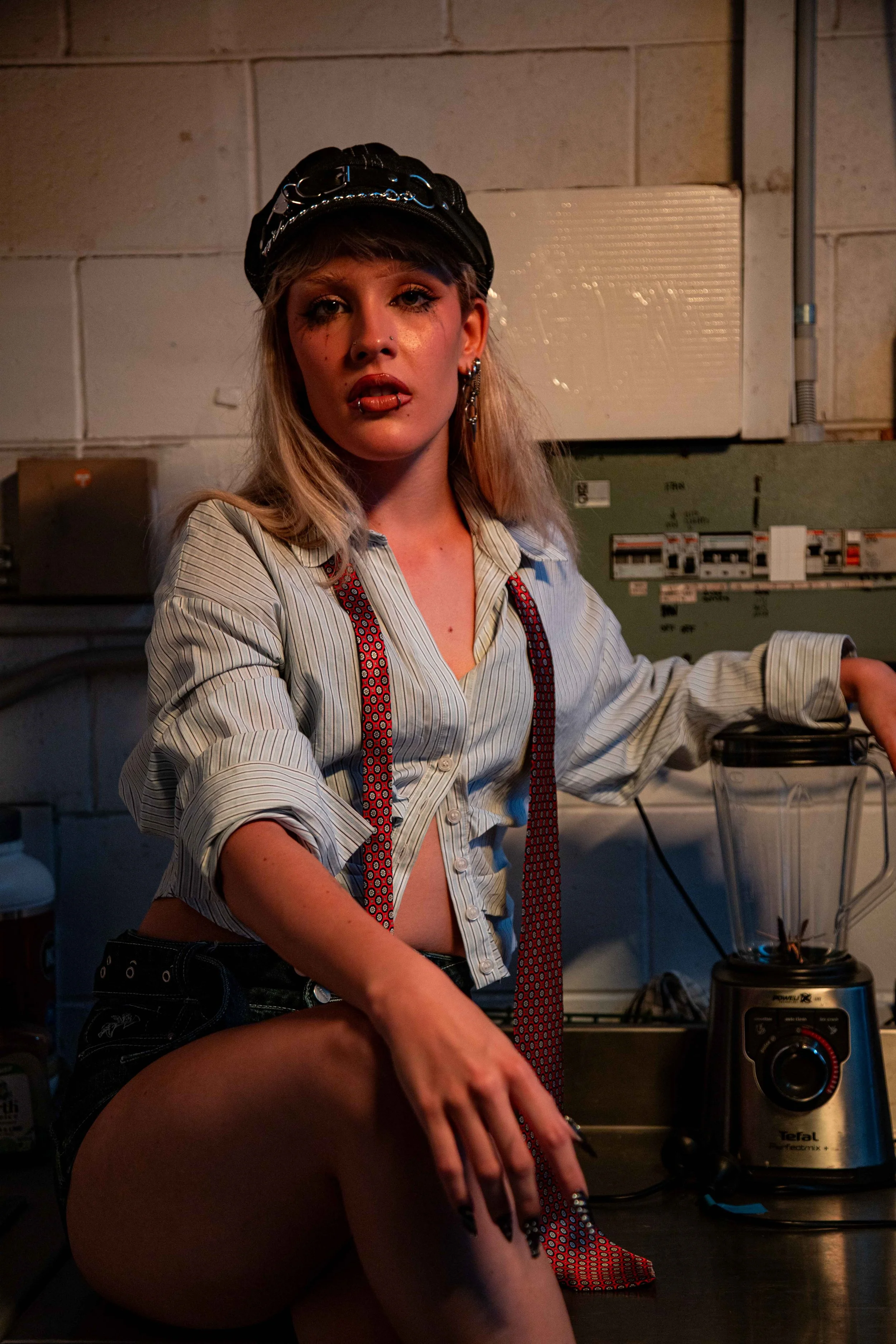 A young woman with blonde hair, wearing a black leather hat, an unbuttoned striped shirt, red suspenders, and shorts, sitting on a workbench in a dimly lit industrial kitchen, with a blender nearby.