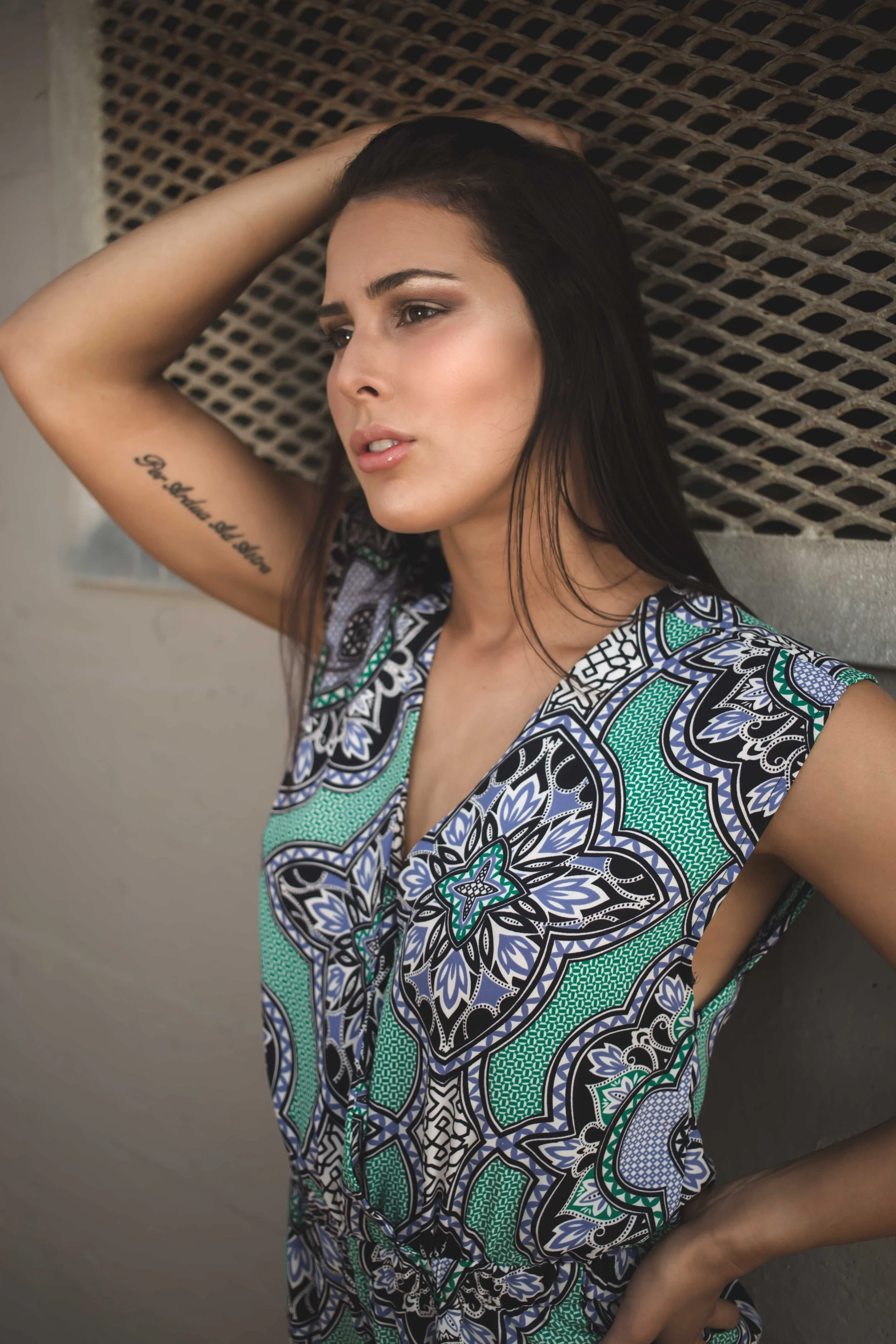 A woman with long dark hair and light skin is posing against a textured wall, wearing a colorful patterned sleeveless top and looking contemplative with one arm raised behind her head.