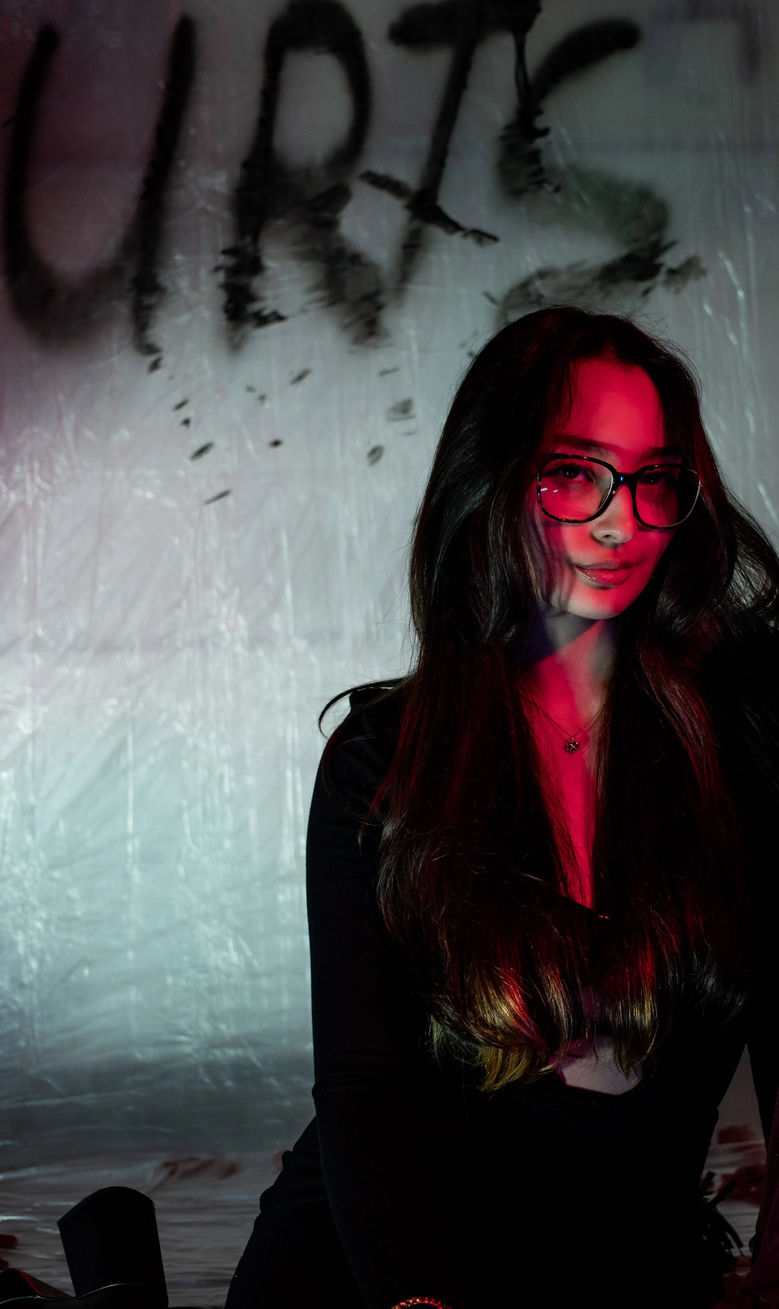 A woman with long dark hair and glasses is illuminated with pink and red lighting, standing in front of a wall with black writing.