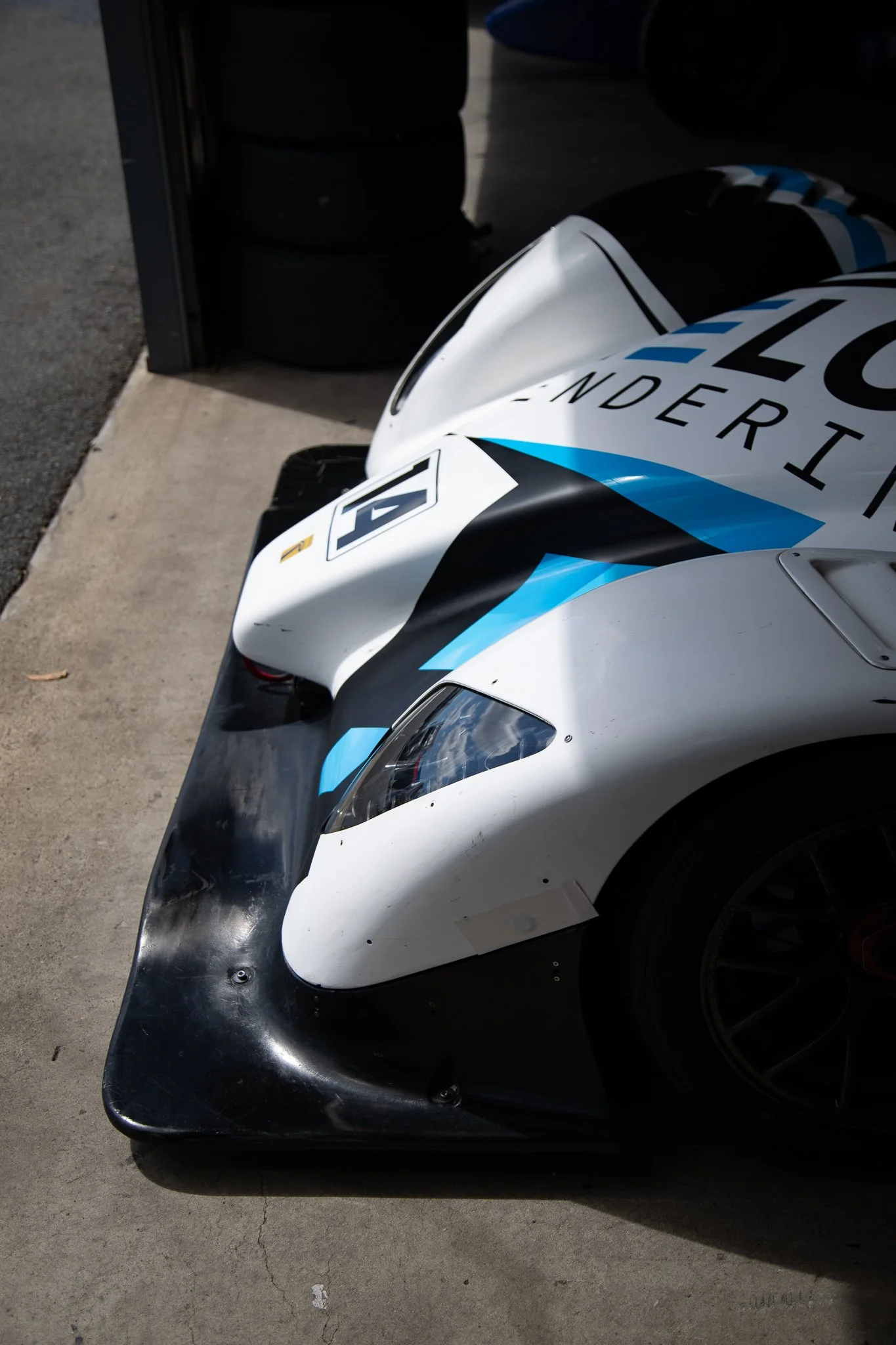 Close-up of a race car's front, showcasing the aerodynamic nose and sleek body, parked on a concrete surface with stacked tires in the background.