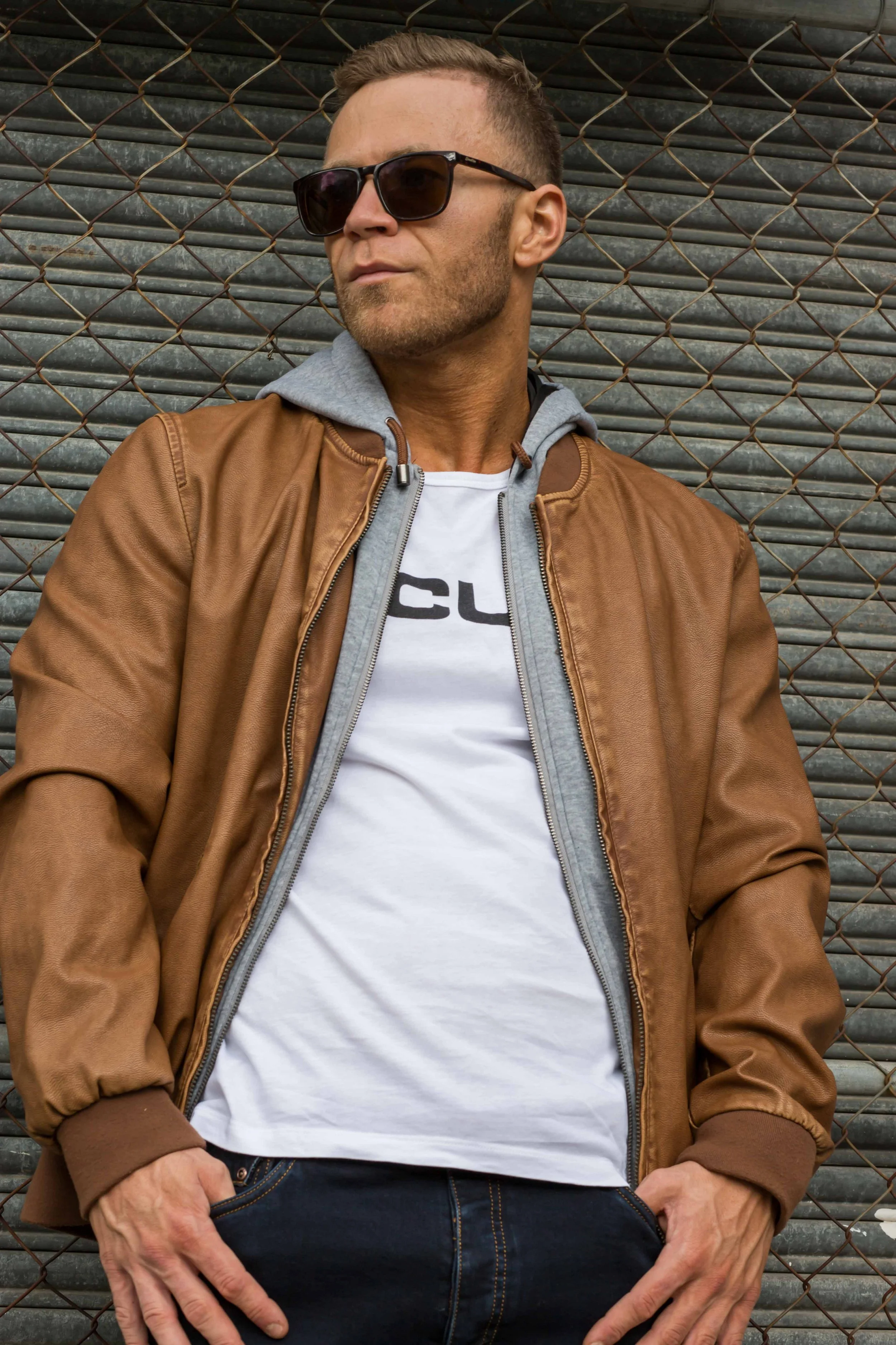 A man wearing sunglasses, a brown leather jacket, a gray hoodie, and a white t-shirt is leaning against a metal chain-link fence.