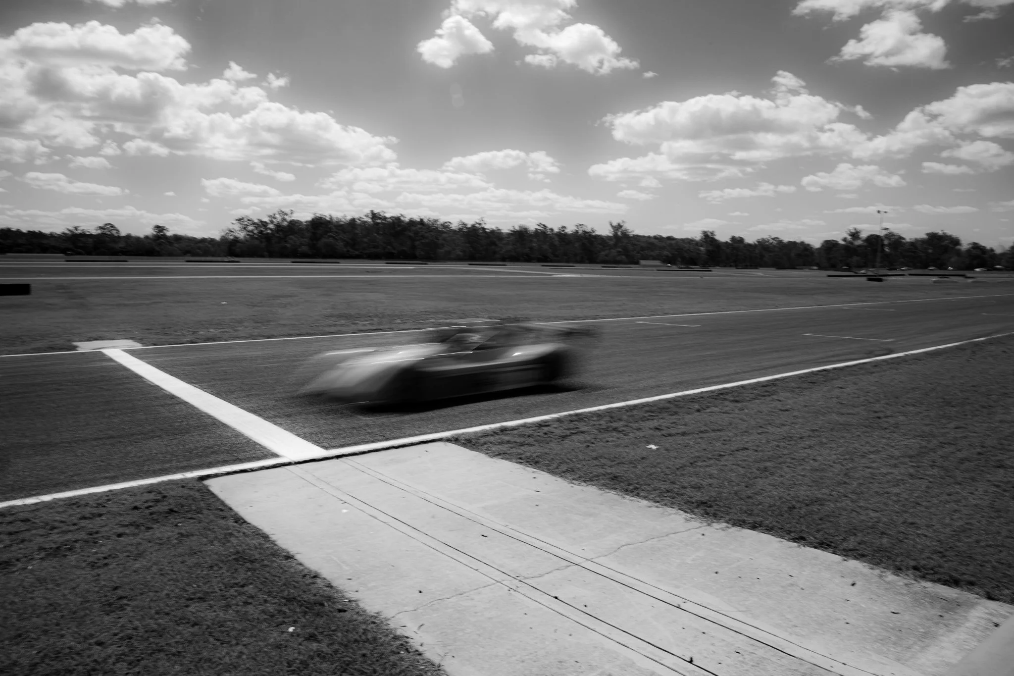 A blurred black sports car on a race track with a grassy field, trees, and partly cloudy sky in the background.