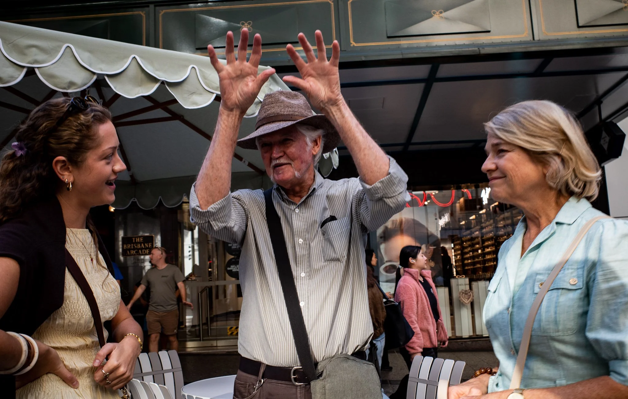 Three people having a conversation outdoors under a canopy. The man in the middle is gesturing with his hands, wearing a striped shirt and hat. The woman on the left has curly hair and sunglasses on her head, wearing a light-colored top and dark jack