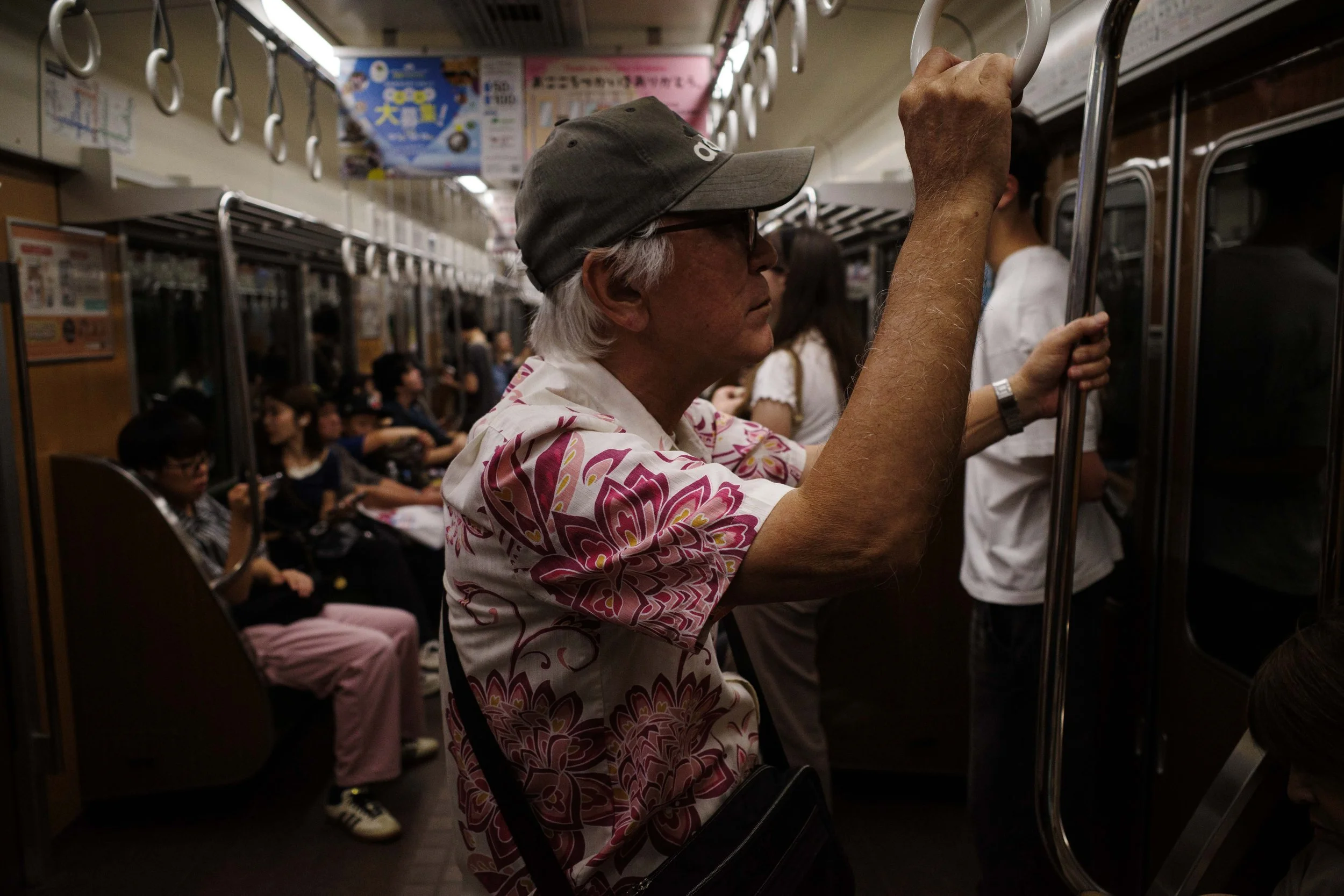 An elderly man with gray hair, glasses, and a gray cap standing inside a crowded subway train, holding onto a handrail.