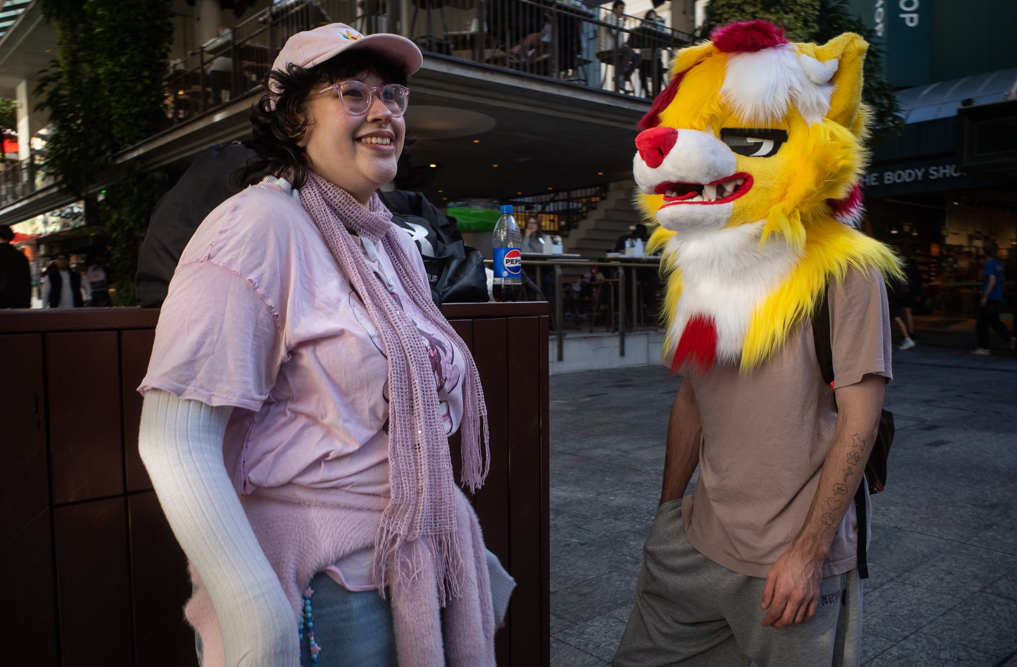 A person with glasses and a pink cap smiling at another person dressed in a fuzzy yellow lion costume with a red mane, in an outdoor urban area with people and stairs in the background.