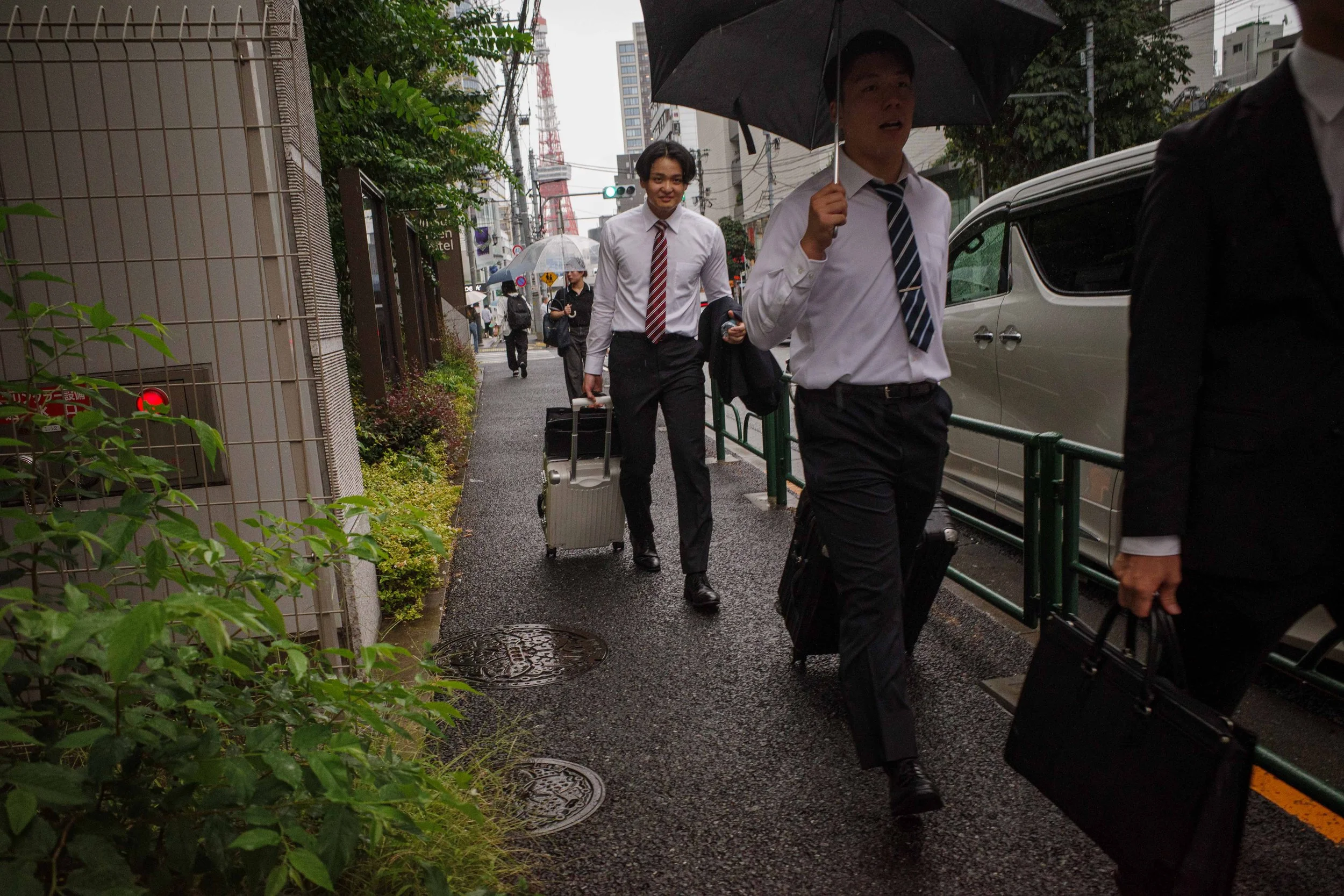 Three businesspeople walking on a rainy city sidewalk with umbrellas, wearing formal suits and carrying briefcases and rolling luggage.