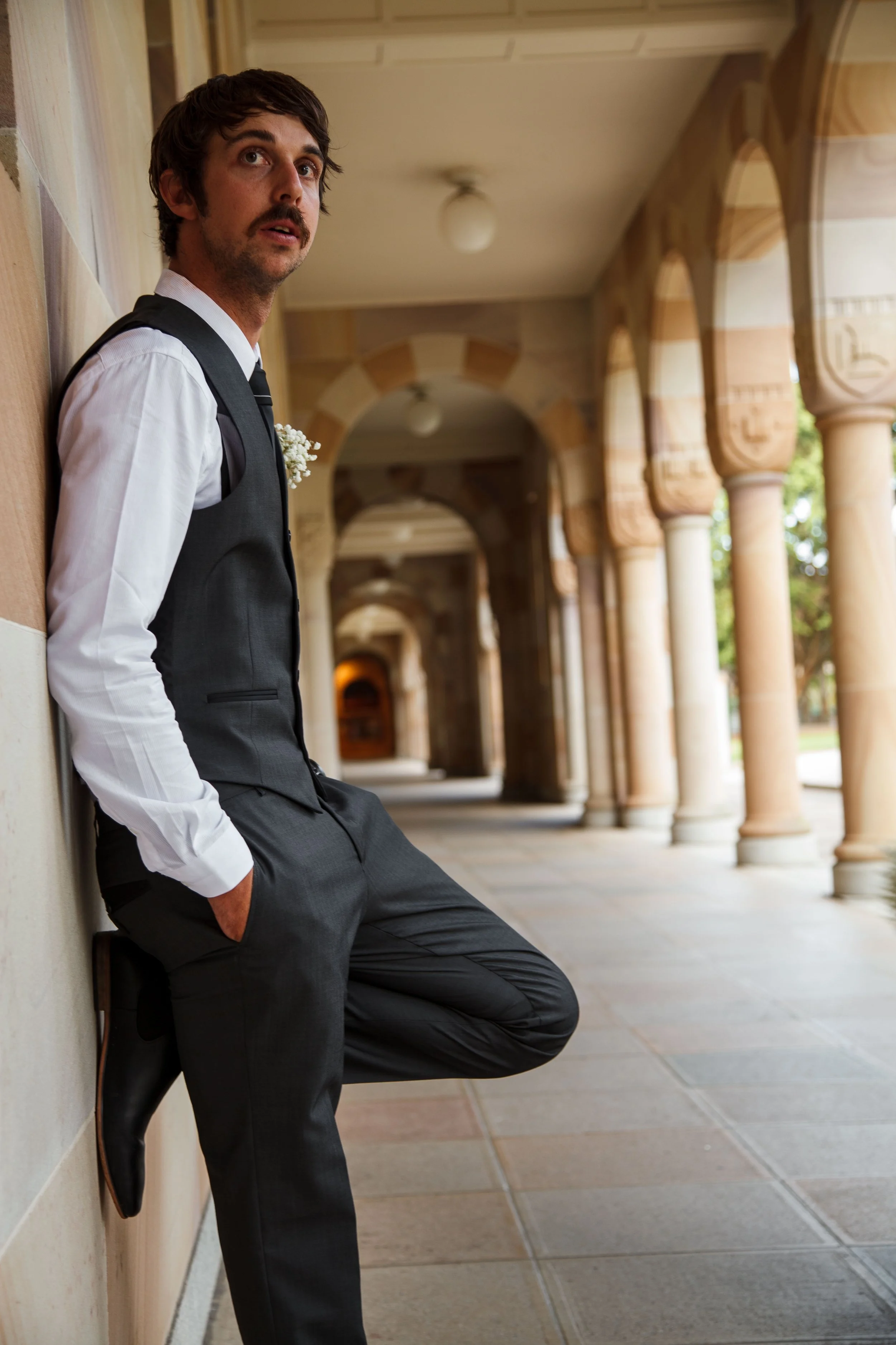A man in formal attire leaning against a wall in an outdoor corridor with arches.