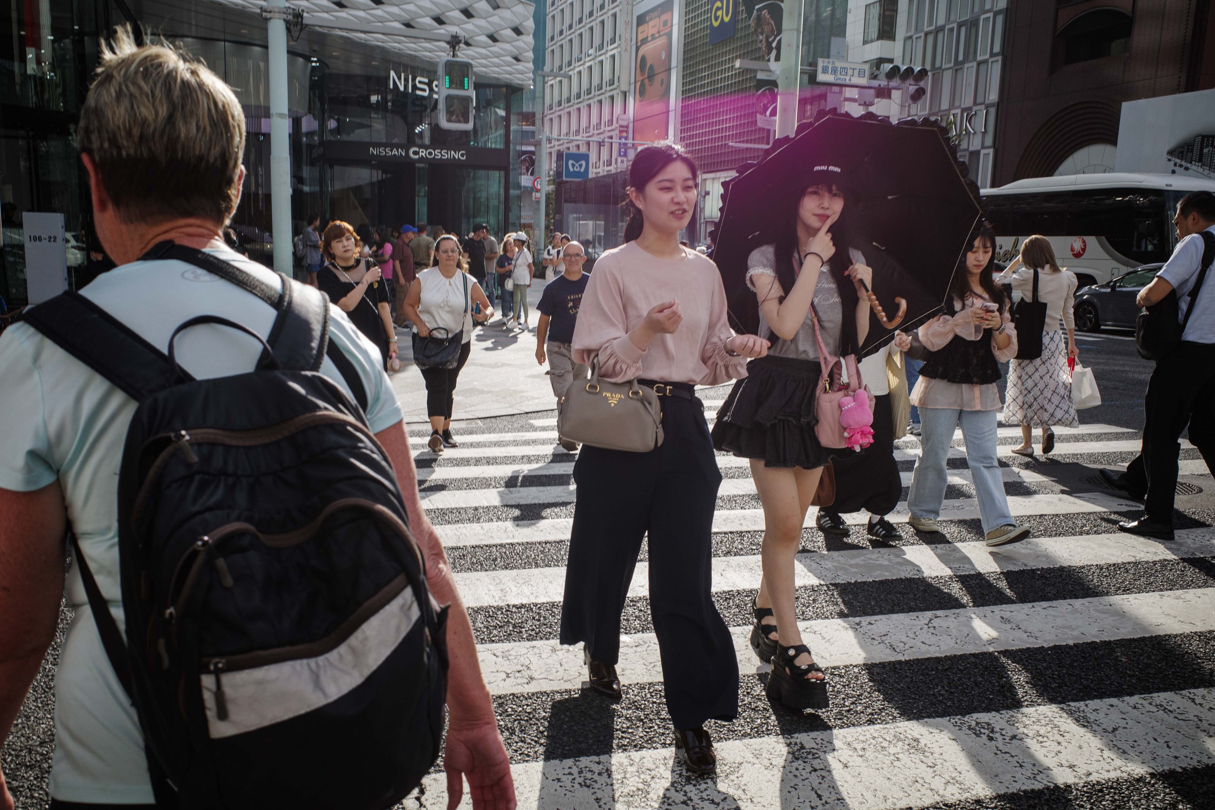 People crossing a busy city street with tall buildings, including a Nissan dealership, in the background. Some individuals are carrying umbrellas, and a woman is looking at her phone.