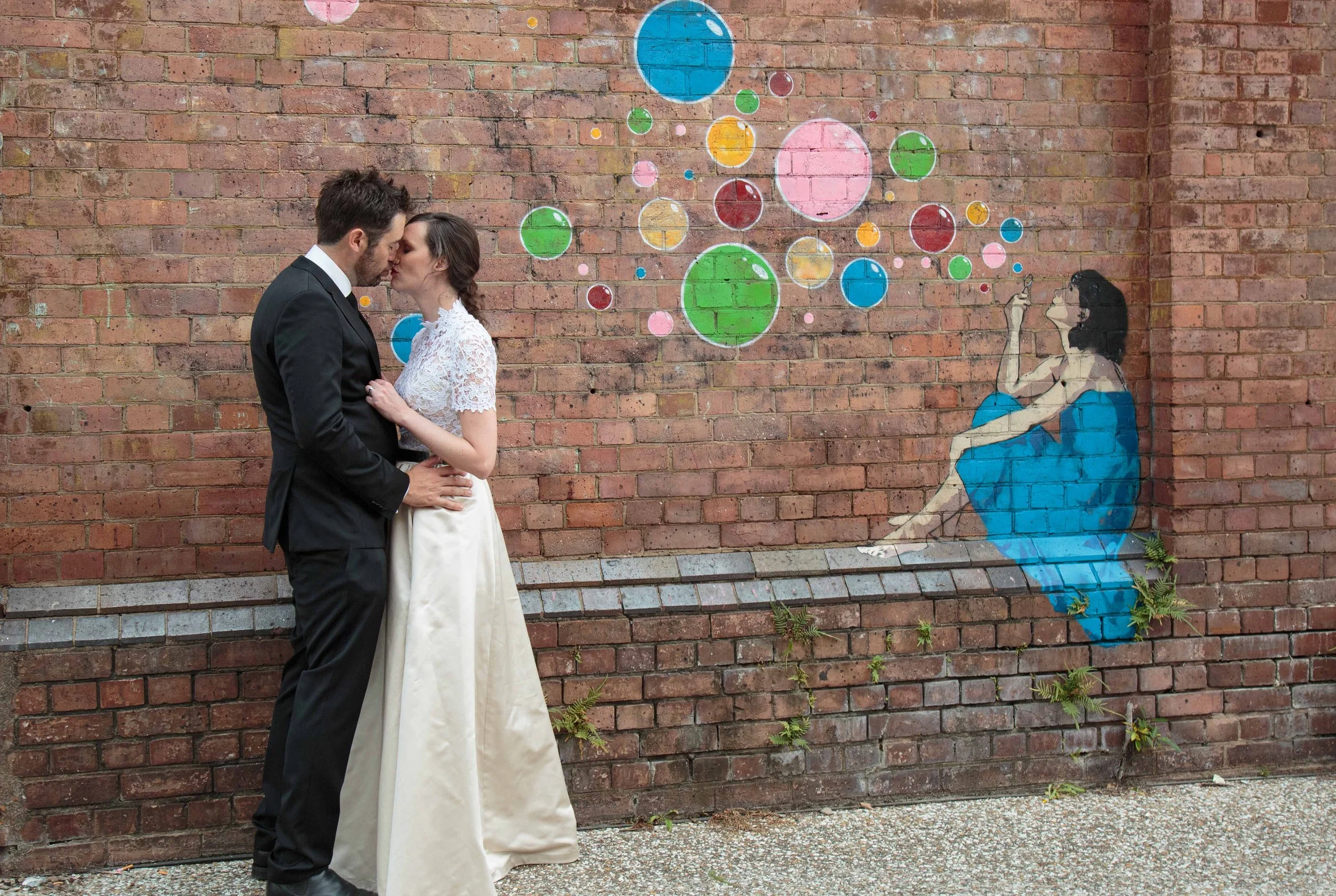 A couple in wedding attire standing close and kissing in front of a graffiti mural on a brick wall. The mural depicts a woman sitting on a bench with her eyes closed, holding a cigarette, with multicolored circles above her.