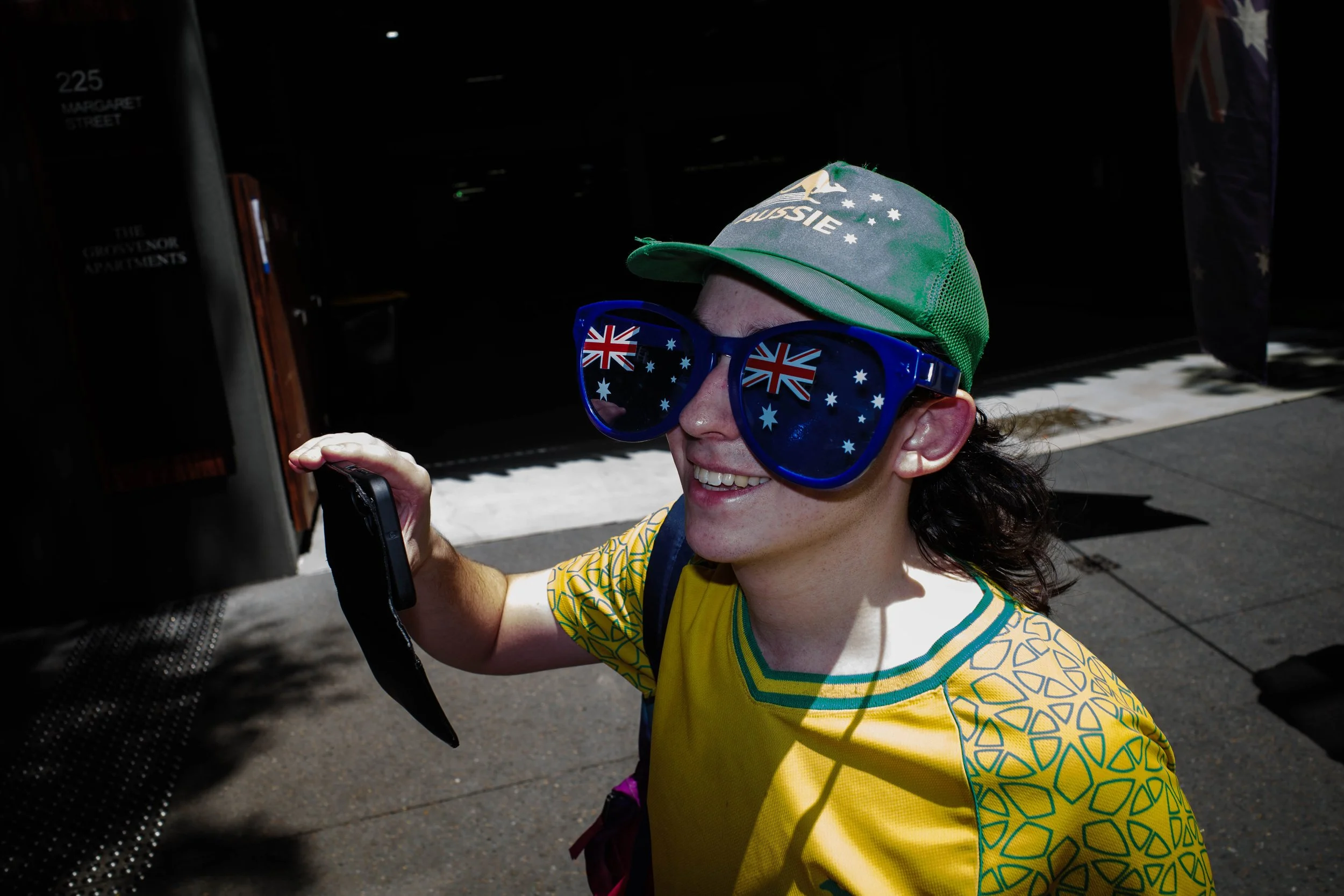 Woman smiling and wearing large sunglasses with Australian flag design, a green cap with Australian flag and writing, yellow patterned shirt, and holding a phone outdoors.