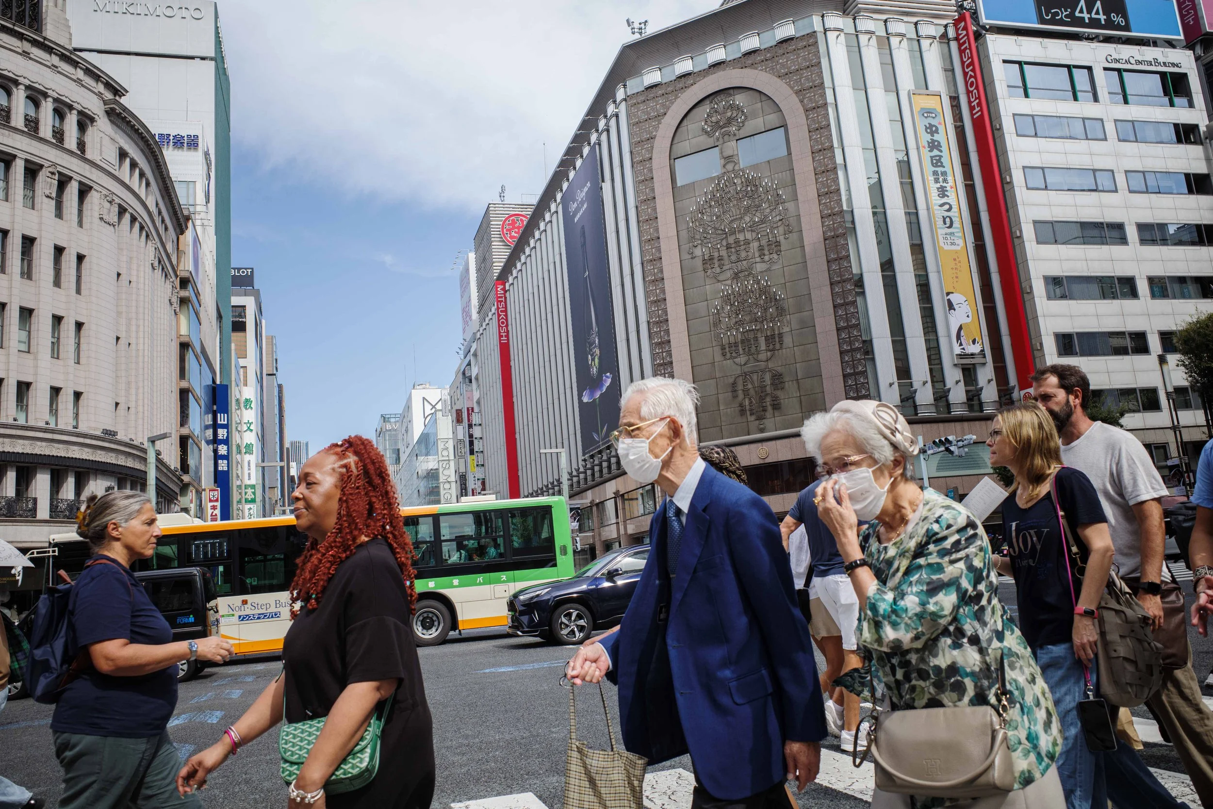 Pedestrians crossing the street in a busy city with tall buildings, some wearing masks, and a green bus in the background.
