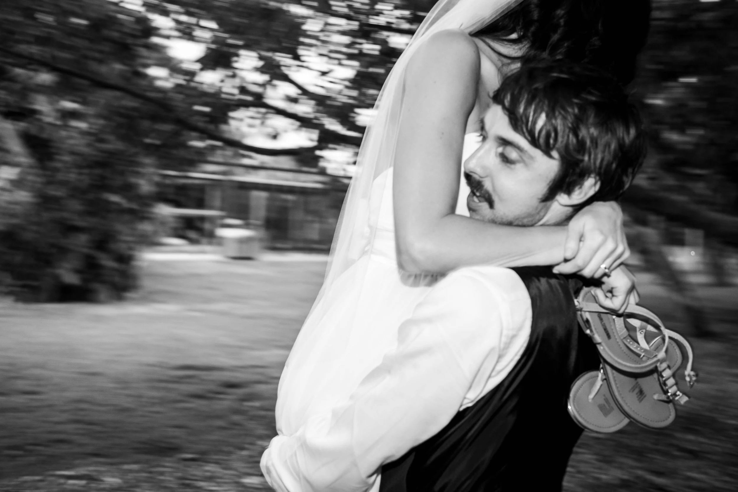 A man in a white shirt carrying a woman in a wedding dress, with the woman hugging him around the neck, in black and white, with blurred background.