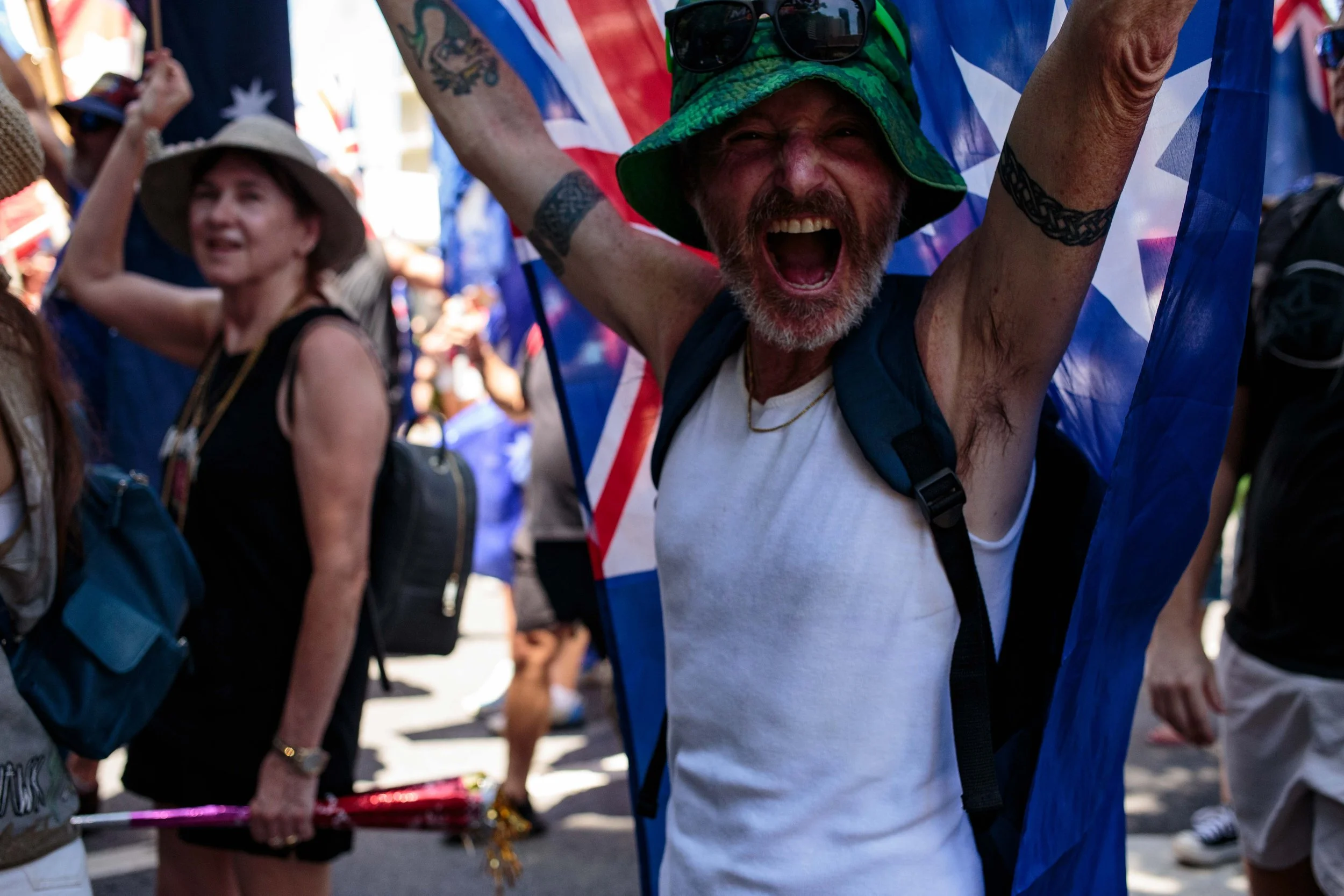 A man wearing a green hat, sunglasses, and a white sleeveless shirt, with a Backpack, is cheering with his arms raised, surrounded by others at a parade or gathering, holding flags and celebrating.