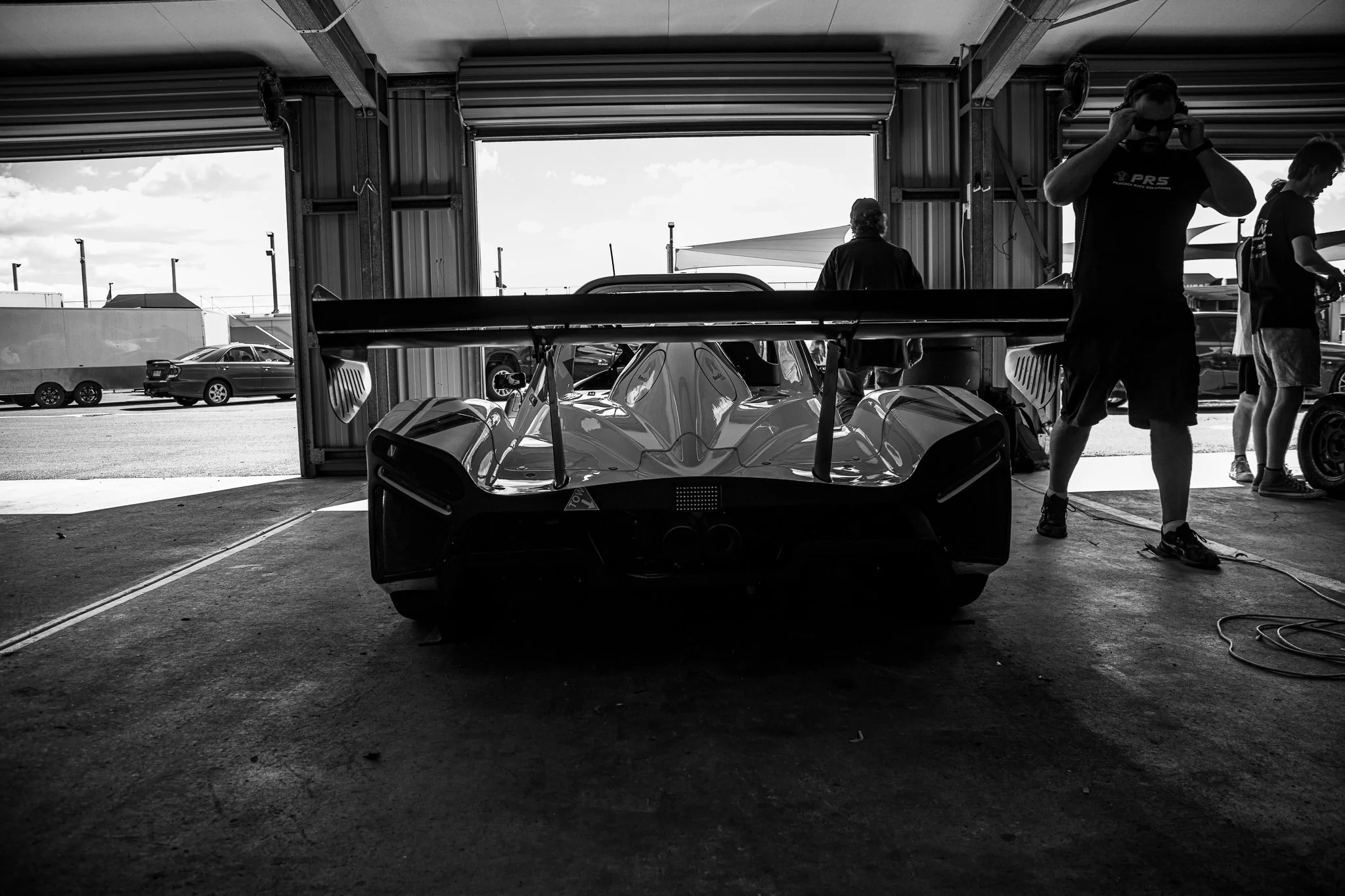 Black and white photo of a race car inside a garage with people around, some working on the car and others standing nearby. The garage door is open, revealing a parking lot with parked cars and a clear sky outside.