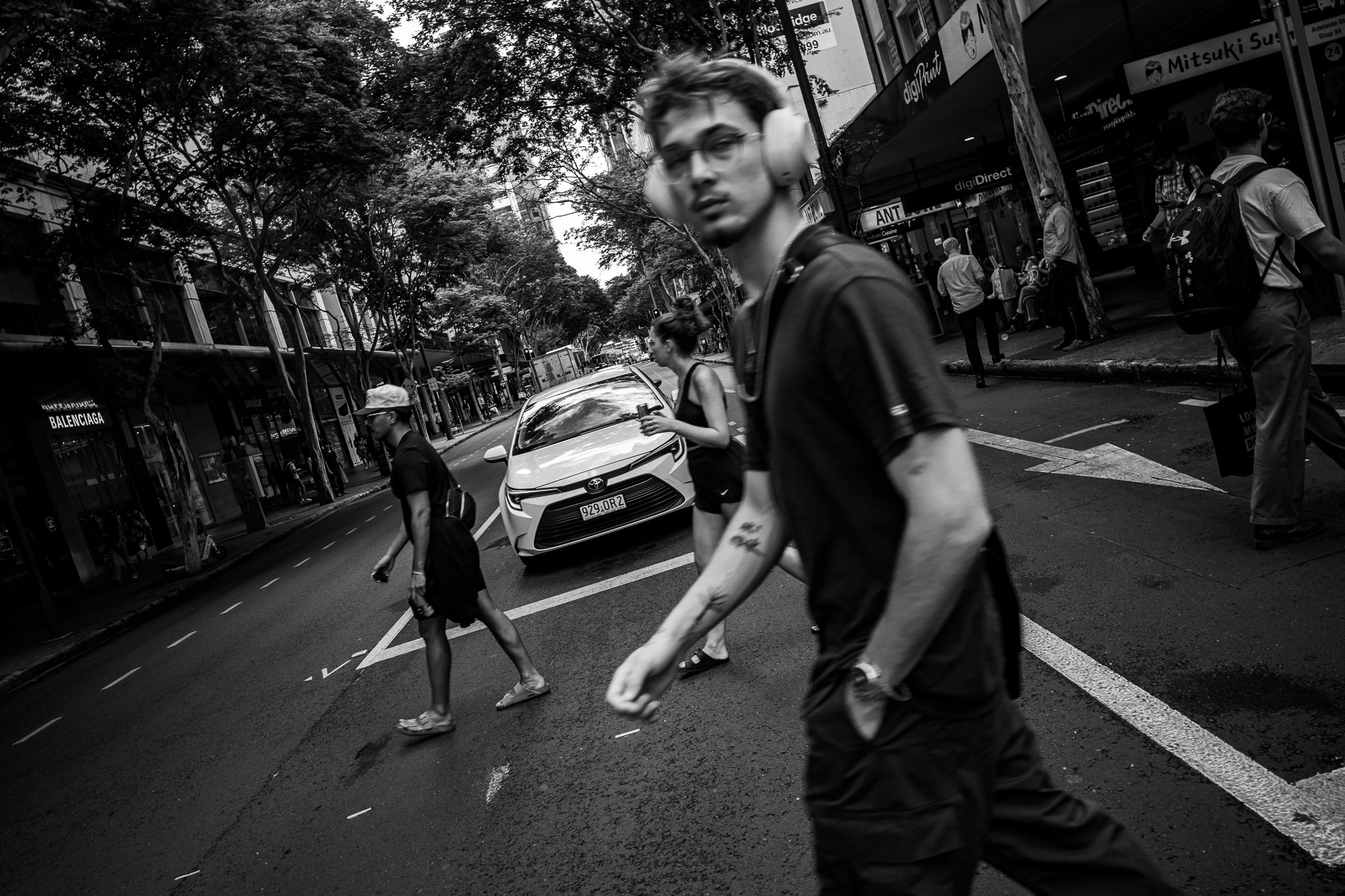 Black and white photo of a city street with pedestrians crossing. A man with glasses and headphones is in the foreground, slightly blurred. Others, including a woman with a coffee, walk on the sidewalk and street.