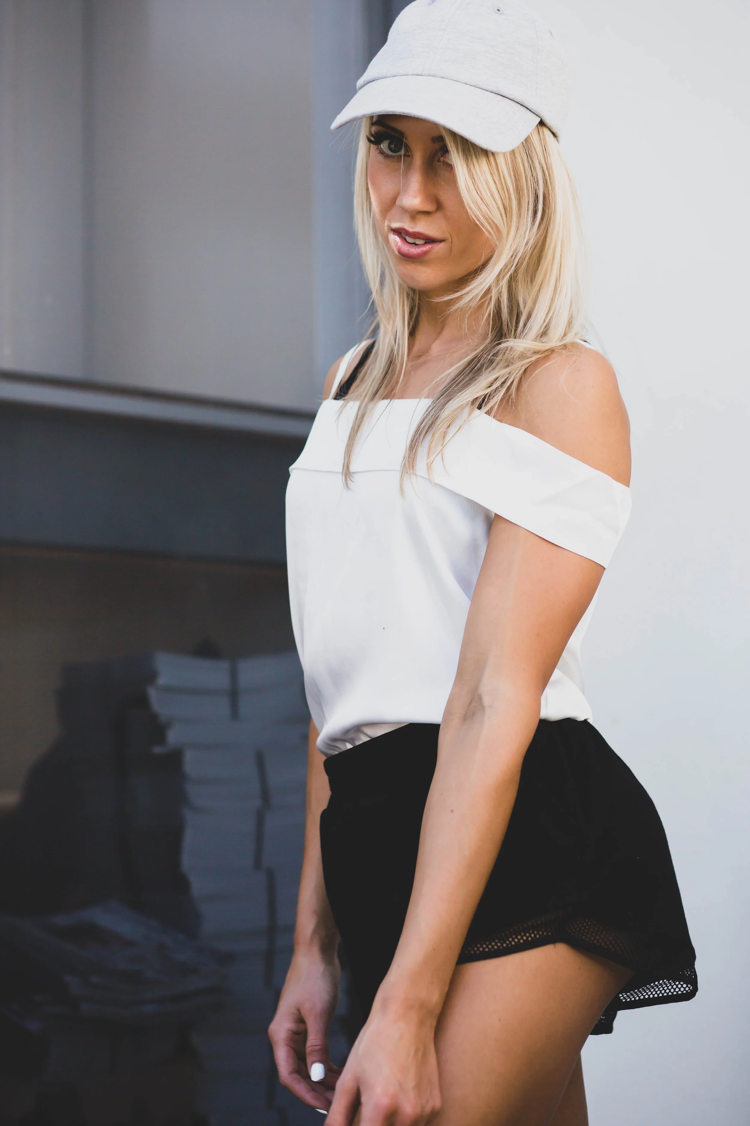 A woman with blonde hair wearing a gray baseball cap, white off-the-shoulder top, and black shorts, standing indoors near a gray wall.