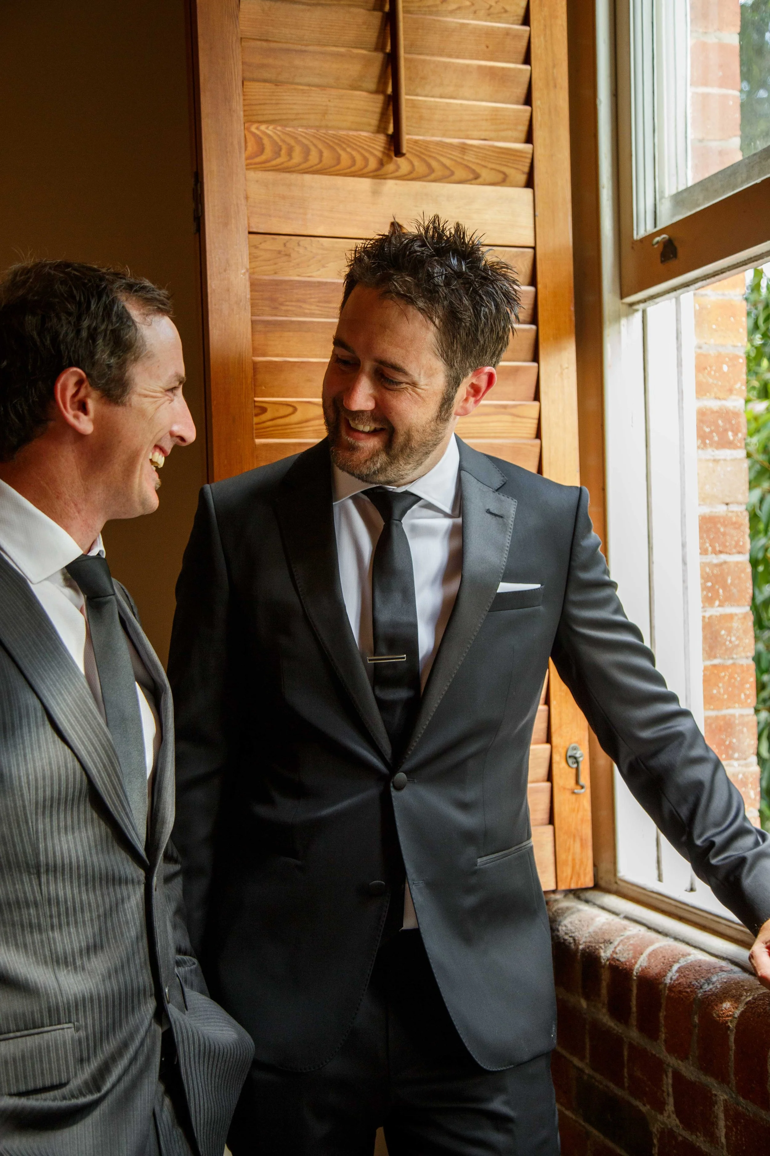 Two men in suits smiling and talking near a window, with a wooden wall and brick ledge behind them.
