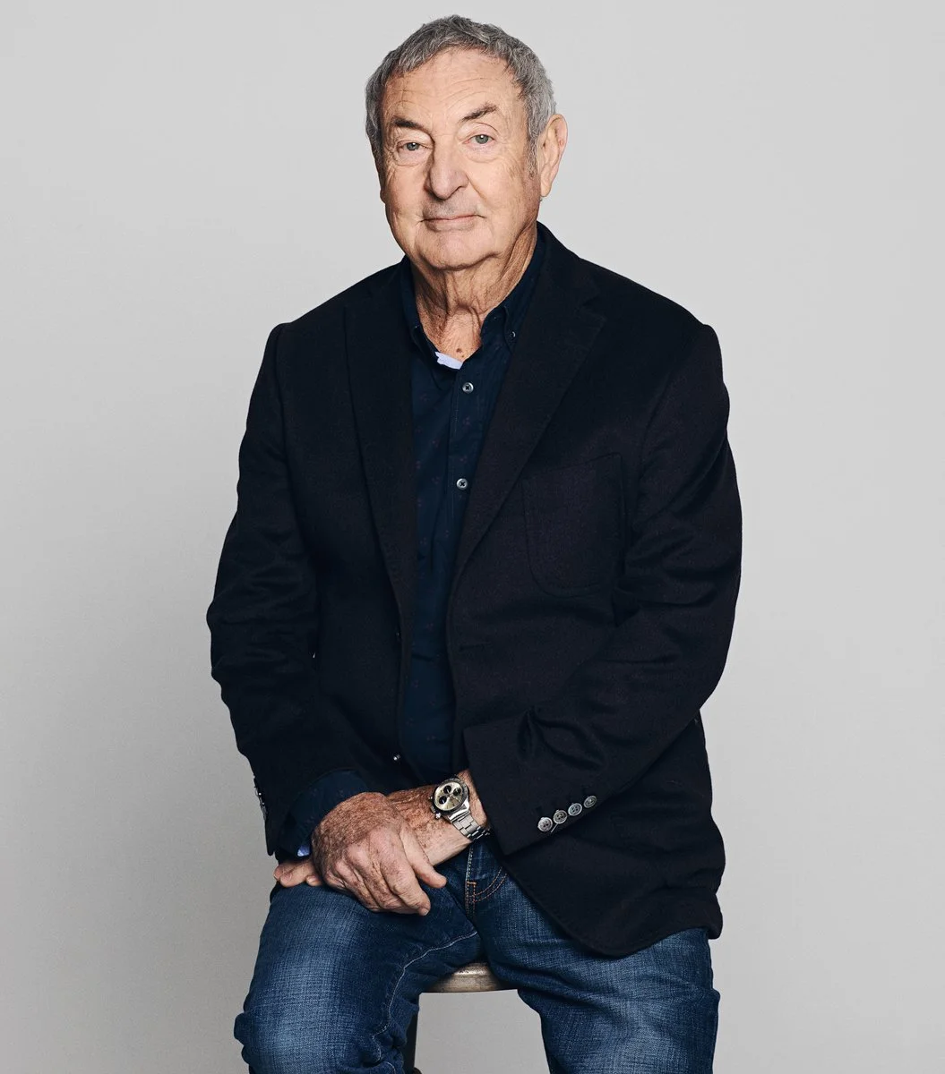 Elderly man in dark blazer and jeans sitting on a stool against a neutral background.