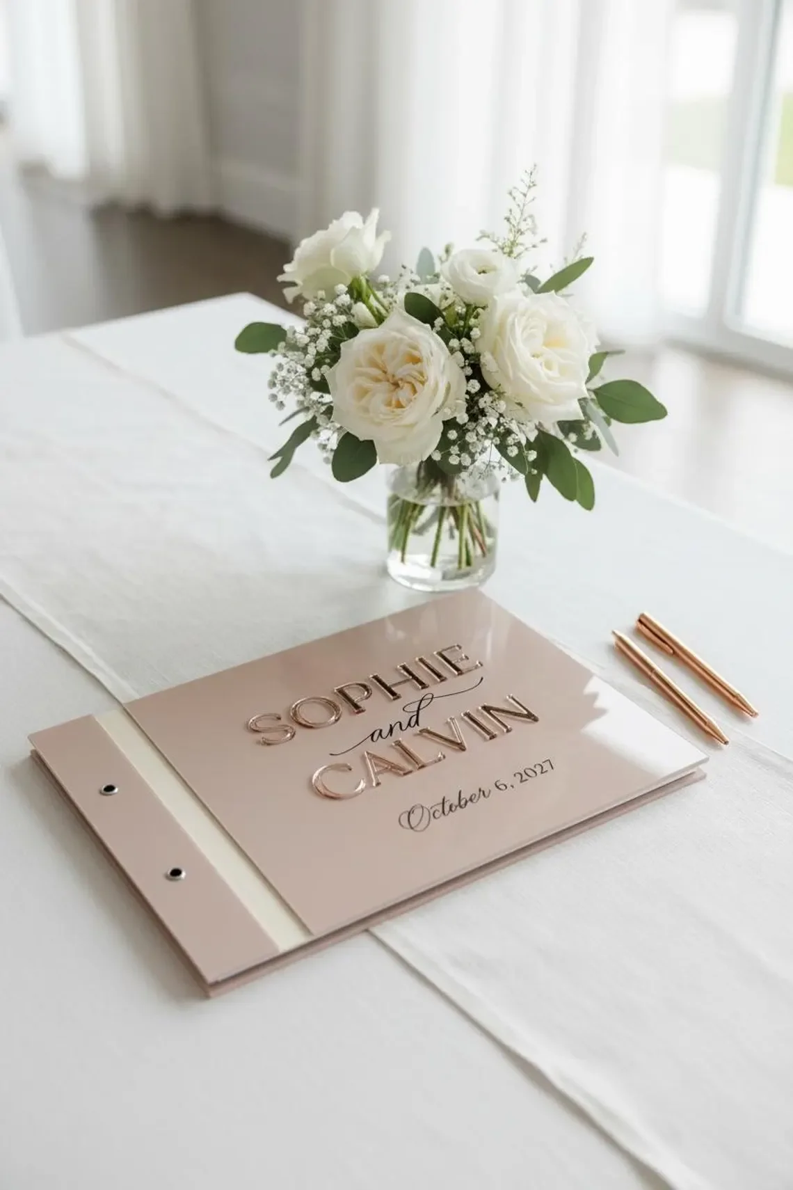A pink guest book with gold lettering that says "Mr & Mrs Clark" and the date "6th of June 2022," placed on a white table with white and beige fabric. Next to the guest book is a white pen. Behind the book, there's a large bouquet of flowers in shades of pink, white, cream, and brown, including roses and other blooms. Also on the table, there are two tall candles in gold holders.
