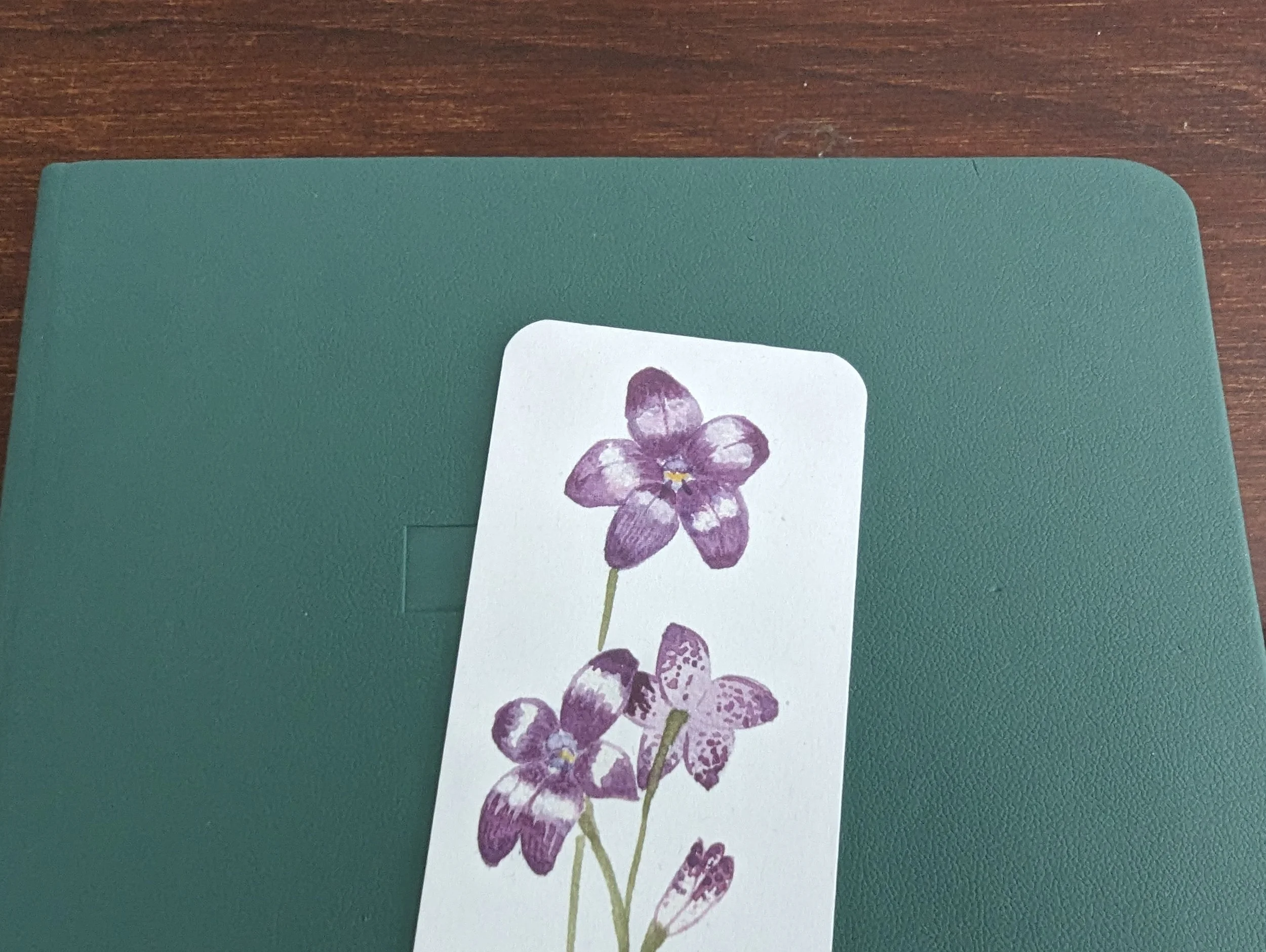 Watercolour purple enamel orchid on white background as a bookmark on a green book on a dark wooden table