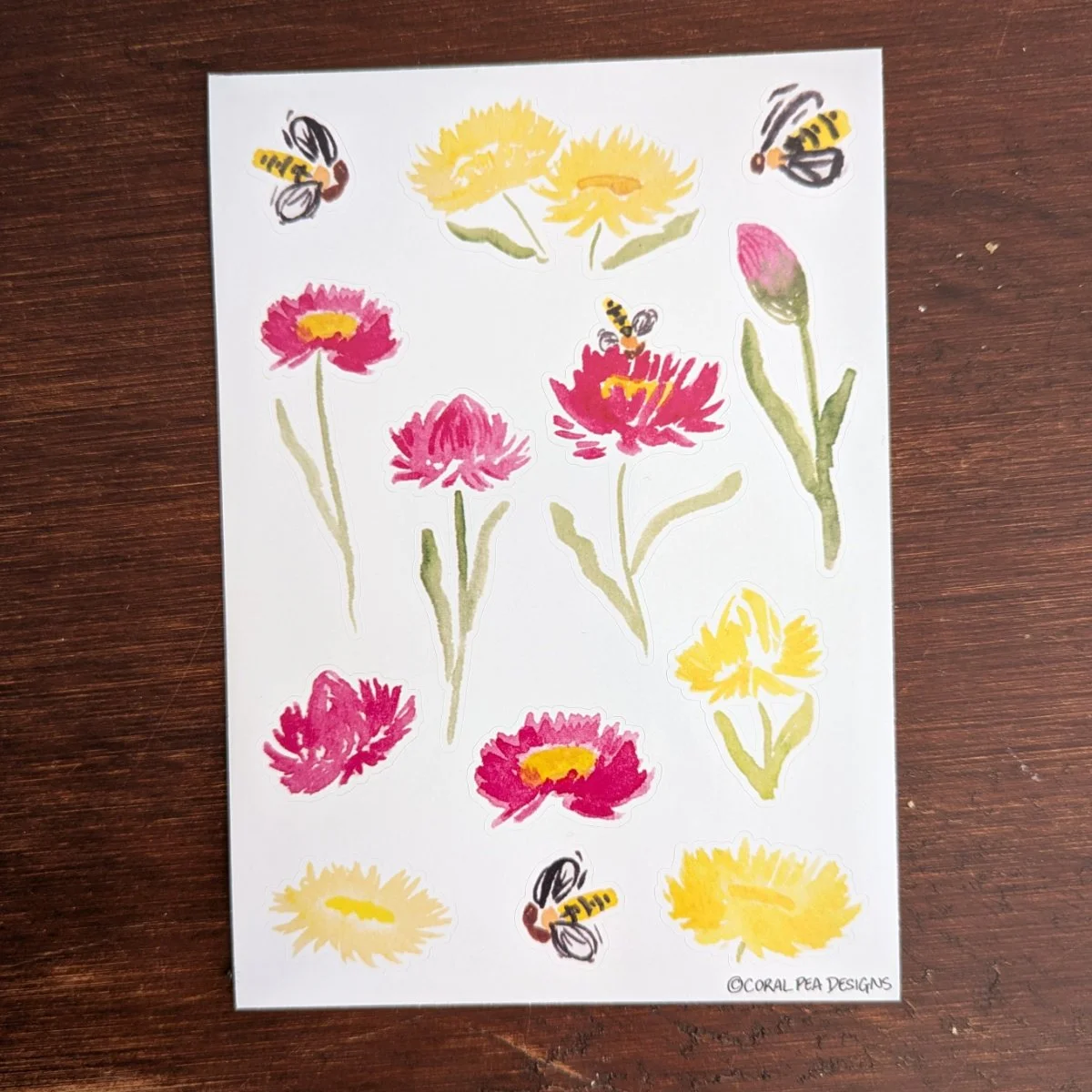 A6 sticker sheet on dark wood table. Stickers are bright yellow and pink daisies, black & yellow hoverflies.