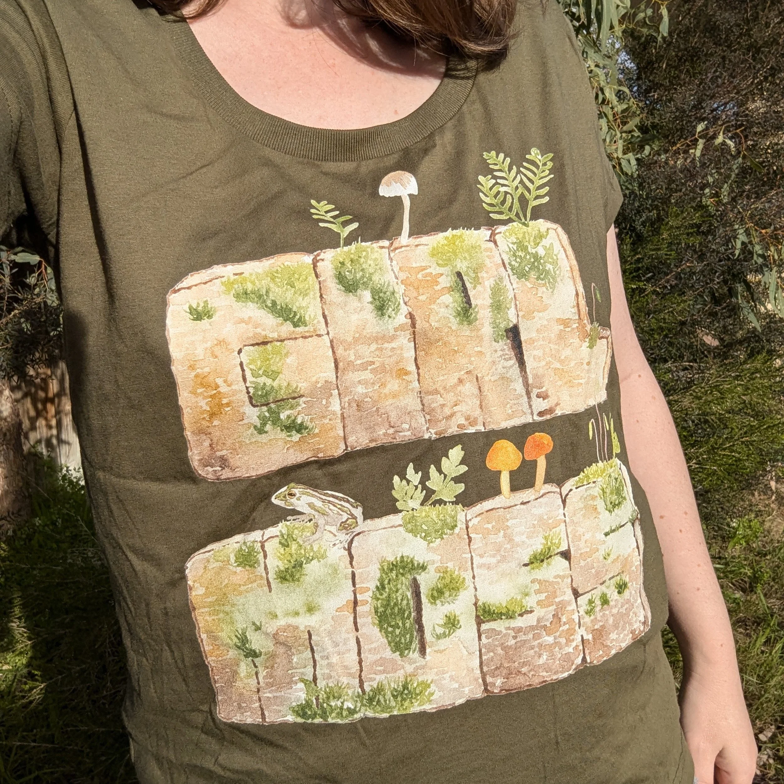 A photo of a woman wearing a green t-shirt which reads "GIRL MOSS". The words are painted in watercolour style like they are limestone rocks, with moss, ferns, frogs and fungi growing all over them.