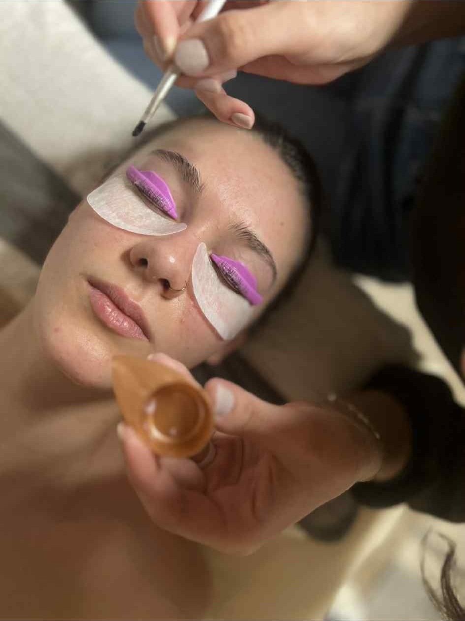 A woman receives lash lift treatment with under-eye pads. An additional person is applying makeup or lash adhesive with a brush.