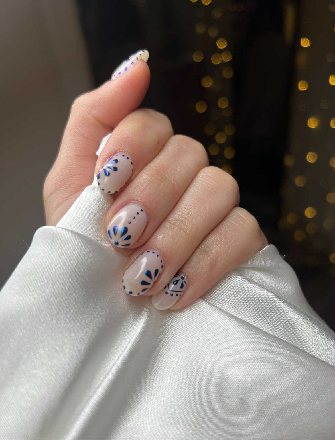 Close-up of a hand with manicured nails painted in a neutral color with blue and black decorative patterns, wearing a white satin garment, against a blurred black background with golden bokeh lights.