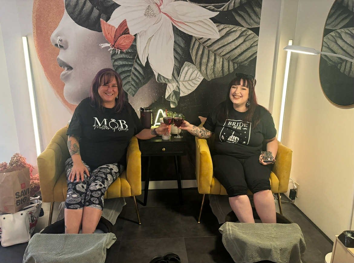 Two women sitting on yellow chairs, clinking glasses of red wine, smiling. Behind them is a large floral mural with a woman's face and white lilies. There are bags and a towel in front of their feet.