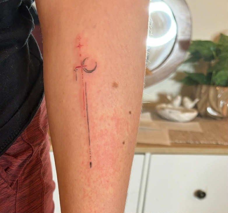 A tattoo on a person's left forearm featuring a black curved line with a small cross below it, a vertical dotted line, and a black dotted line at the bottom, with a light red background around the area.