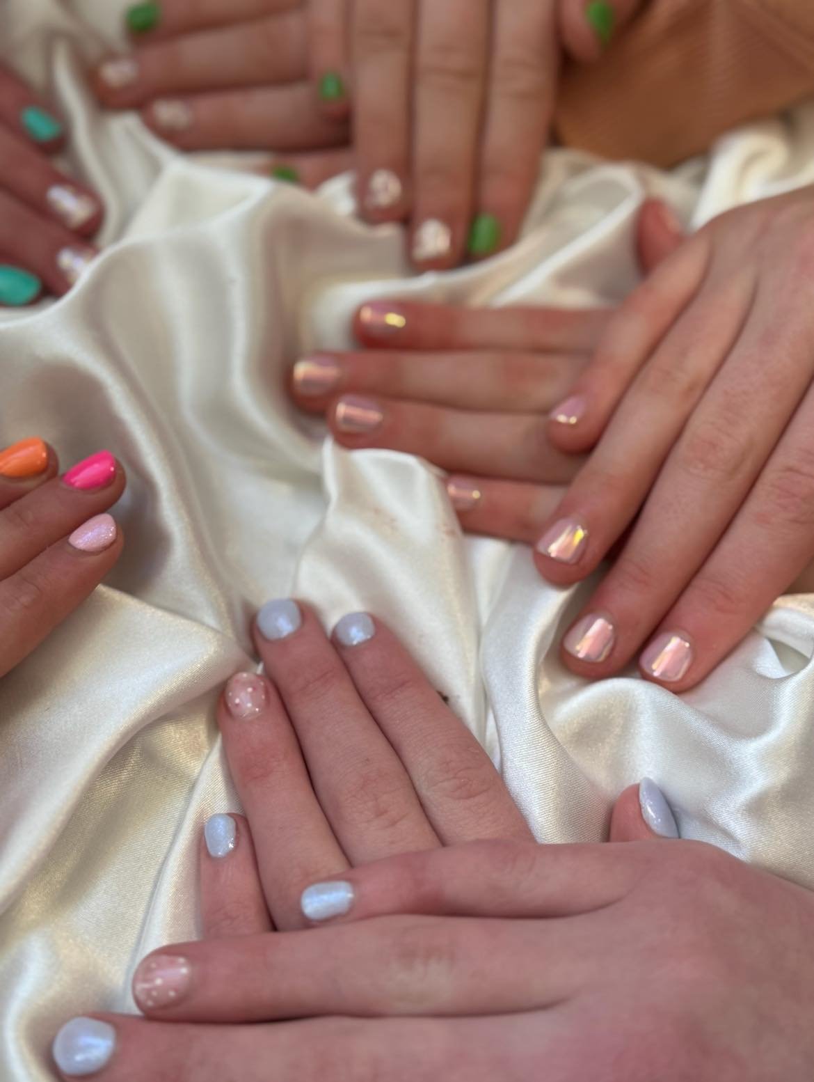 Multiple hands with colorful, pastel, and neutral nail polish resting on cream-colored satin fabric.