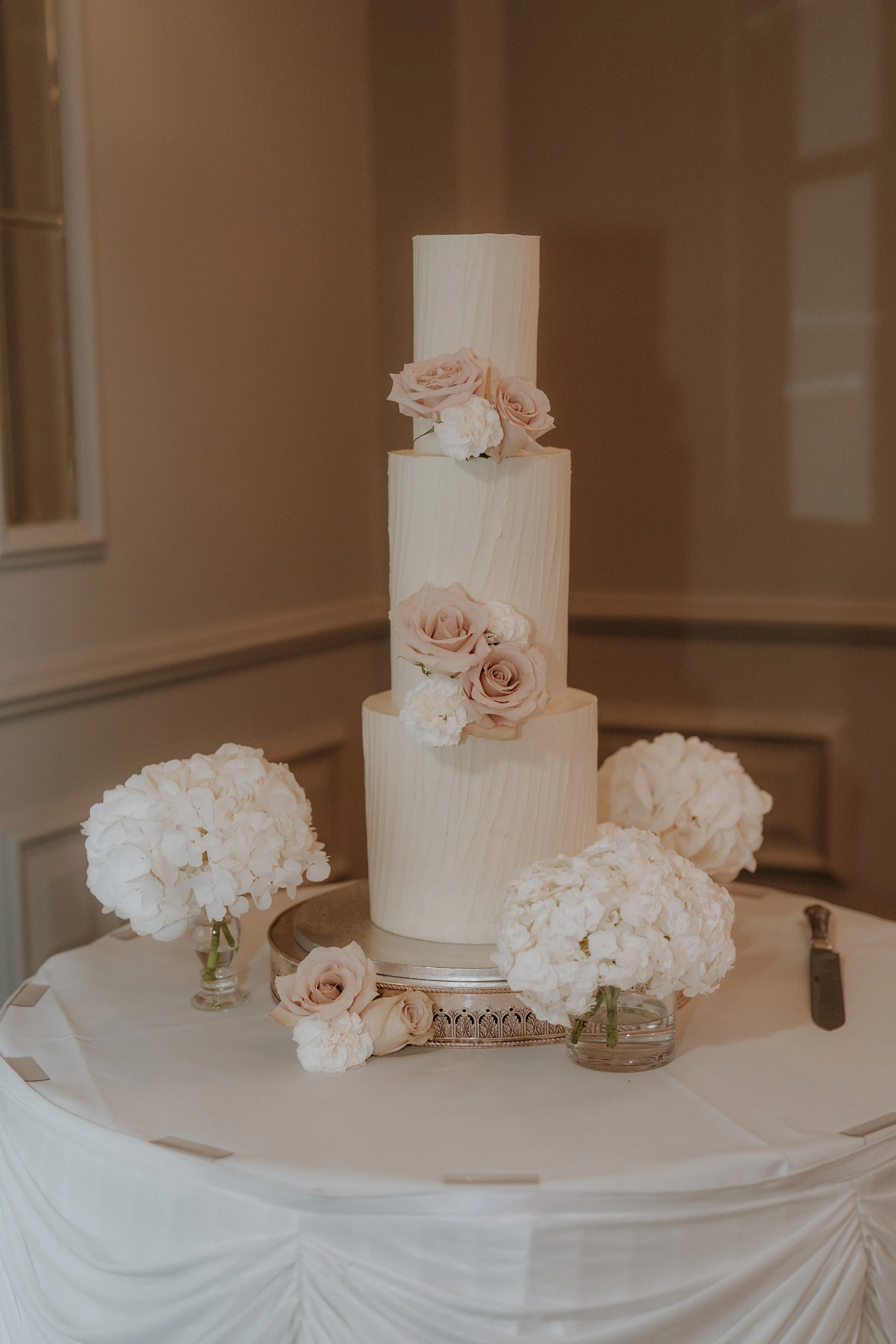 A multi-tiered wedding cake decorated with pale pink roses and white flowers, surrounded by matching flower arrangements on a table with white tablecloth at Sopwell House.