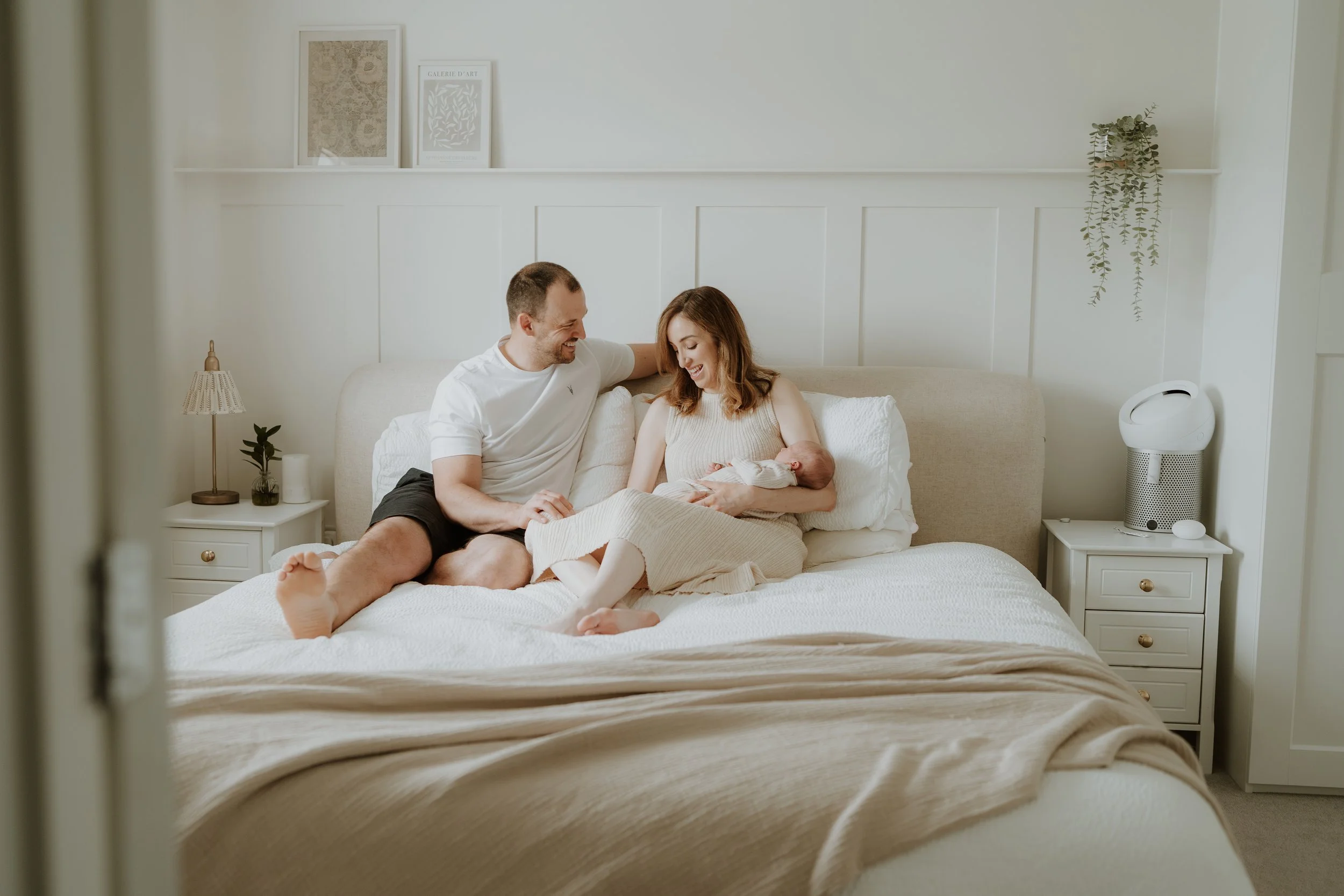 A happy family in a well-lit bedroom, with a woman holding a newborn baby, a man sitting beside her, all smiling.
