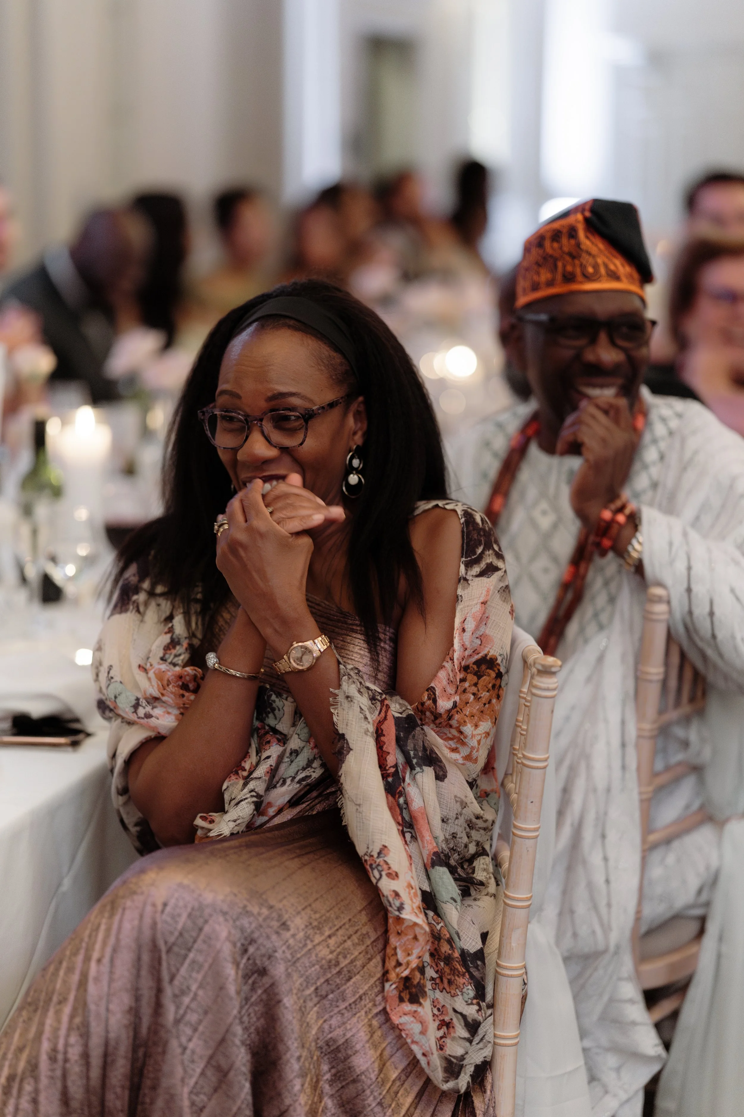 A woman and a man sitting at a banquet, smiling and laughing, dressed in elegant clothing, with the woman covering her mouth in amusement during speeches at Sopwell House.