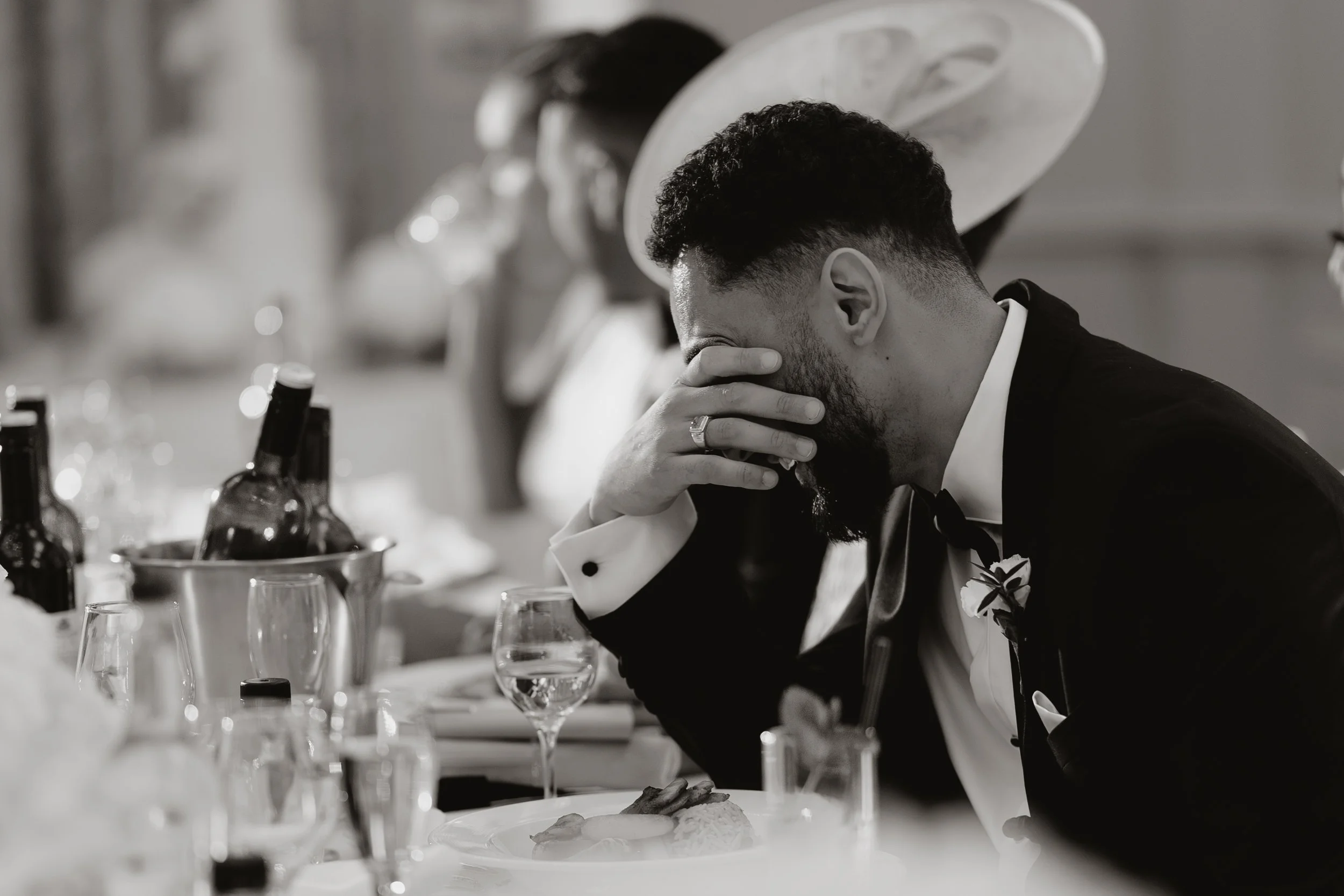 Groom in a tuxedo sits at a table with his hand covering his face, appearing emotional or upset, during speeches at Sopwell House.
