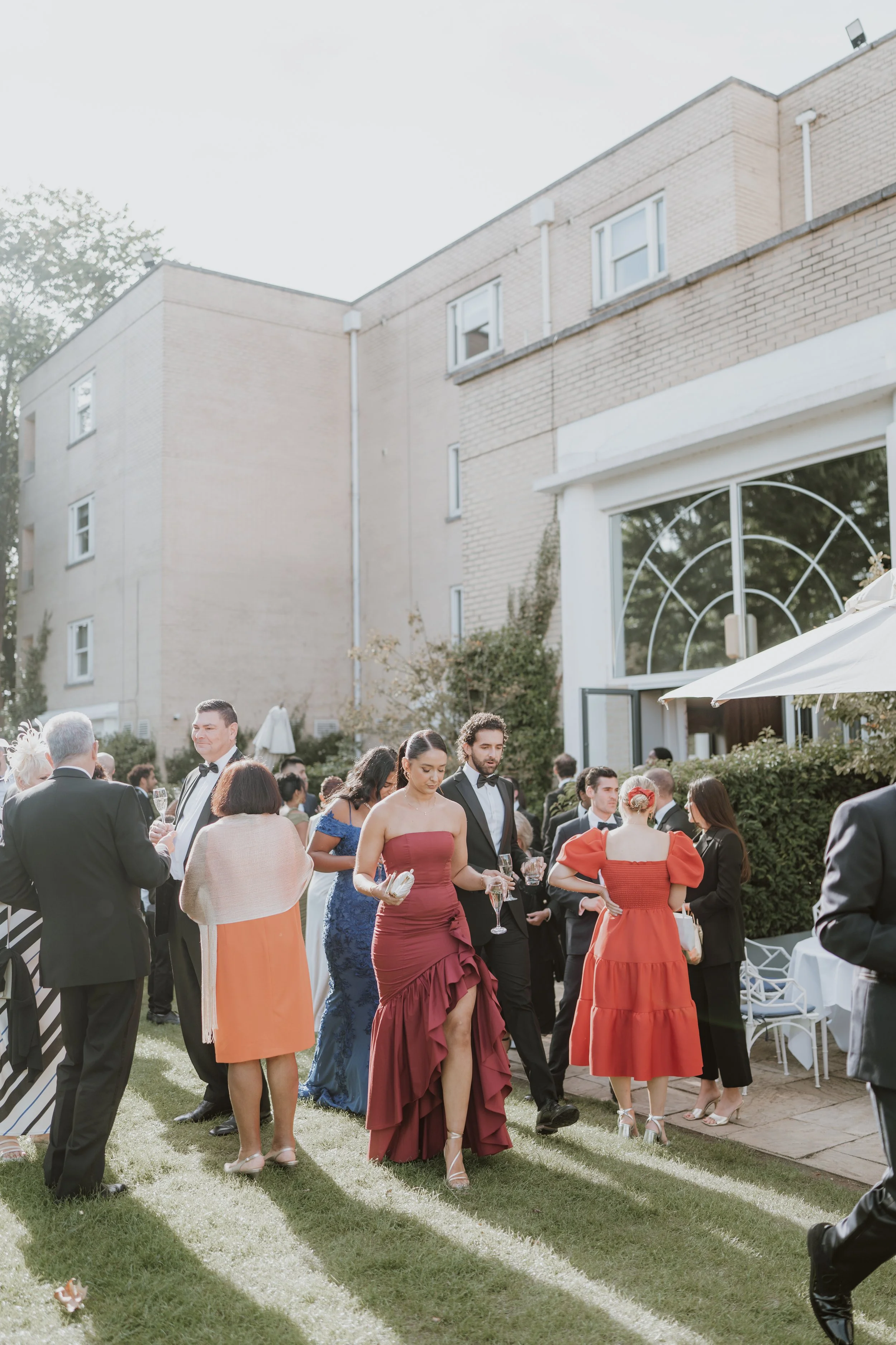 Guests at an outdoor wedding reception on a grassy lawn at Sopwell House, wearing formal attire, some with drinks in hand, near a white building with large windows and an umbrella.