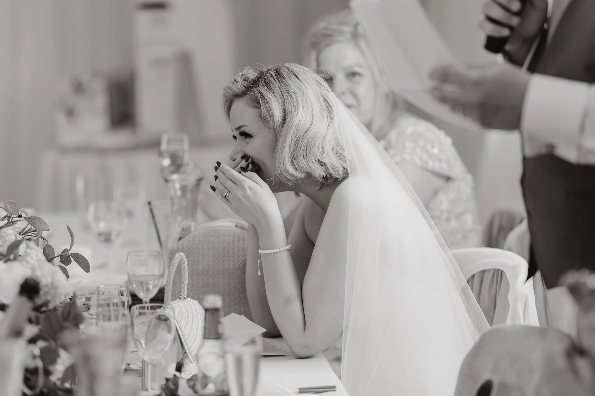 A bride with blonde hair in a bob style, wearing a wedding dress and veil, is sitting at a table, smiling and covering her mouth with her hand. She has a pearl bracelet and ring on her fingers. In the background, another woman, likely a guest, and a