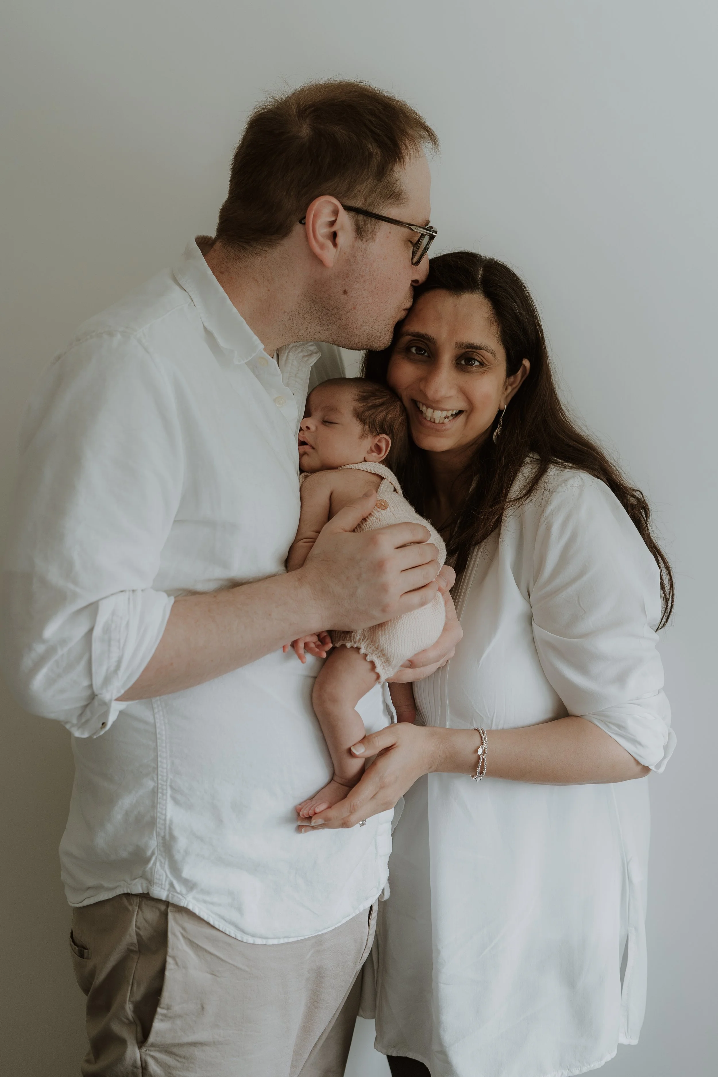 Family of three: a man kisses a woman on the forehead while holding a newborn baby, all dressed in white against a plain wall.