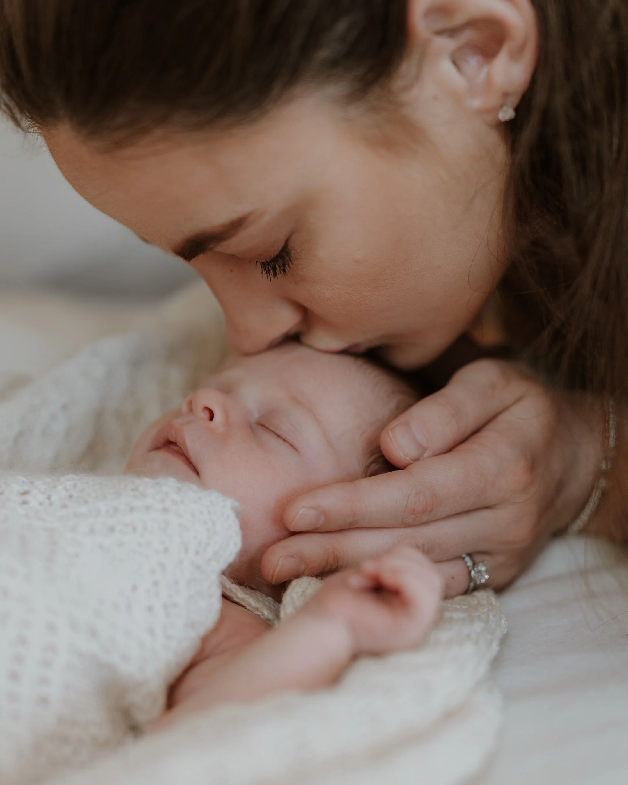 Woman gently kissing sleeping baby on the forehead, cradling baby's head with one hand, wearing a ring and earrings.