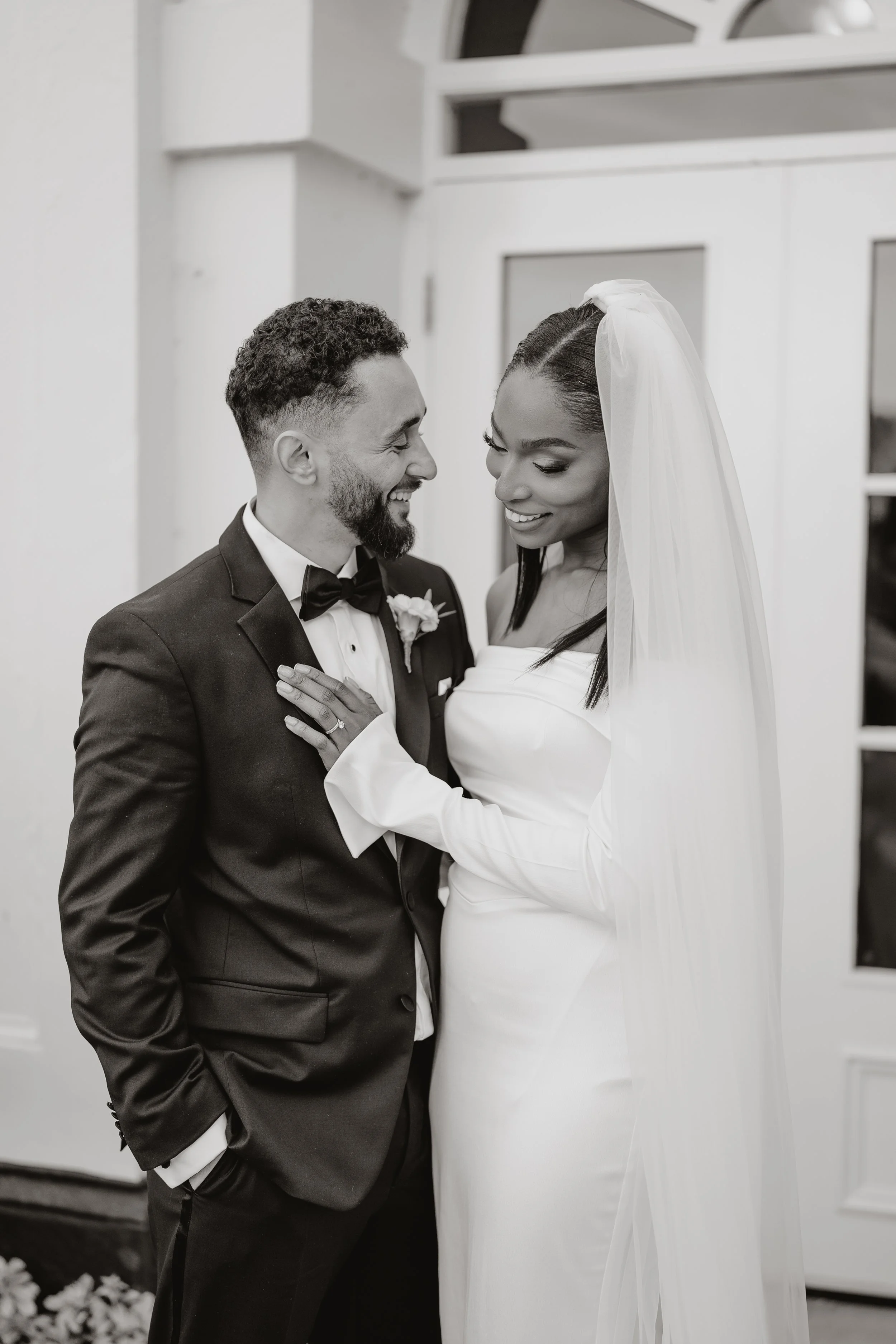 A black-and-white photo of a bride and groom sharing a happy moment during their wedding at Sopwell House. The groom is in a tuxedo with a bow tie, and the bride is in a long white dress with a veil. They are smiling and looking at each other affecti