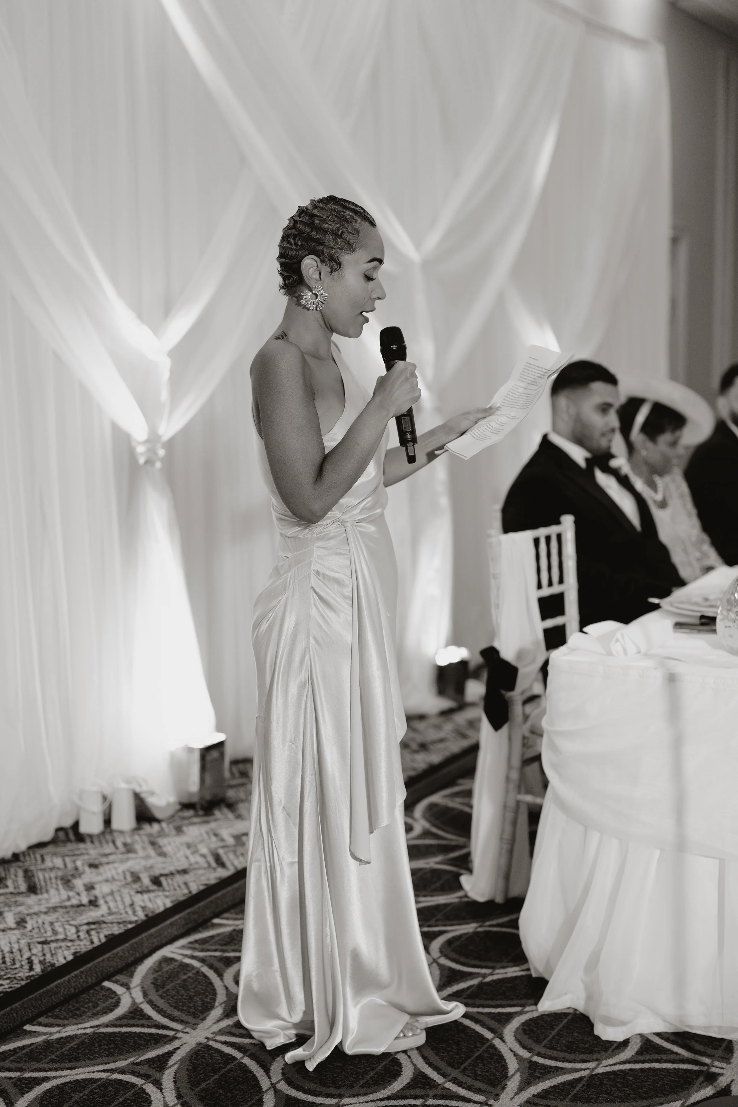 A bridesmaid in a satin dress giving a speech at a wedding at Sopwell House, holding a microphone and reading from a paper, with seated guests in formal attire in the background.