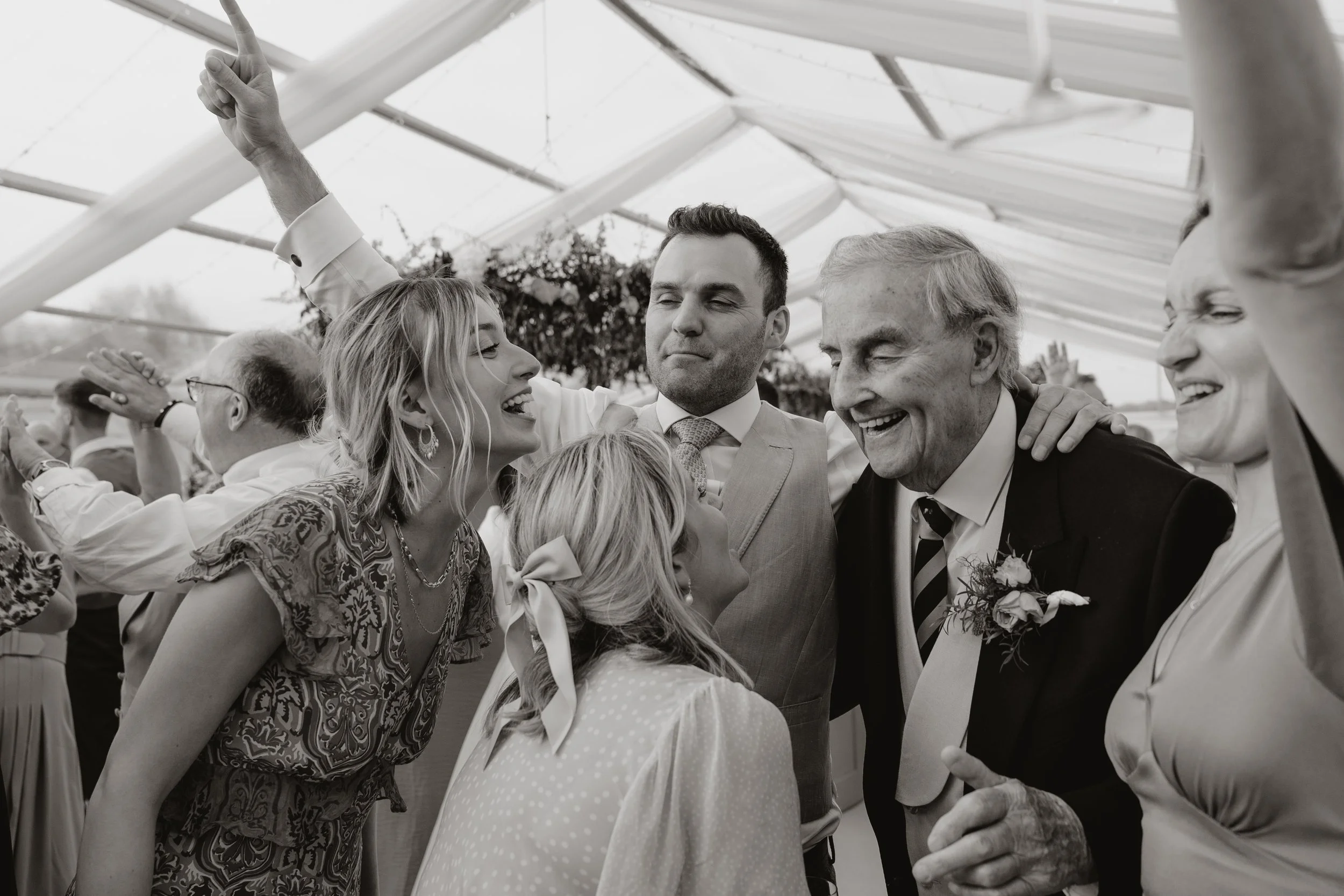 Generational adults celebrating and smiling at a wedding reception, with some dancing and in a large glass greenhouse or conservatory at Eltham Hall.