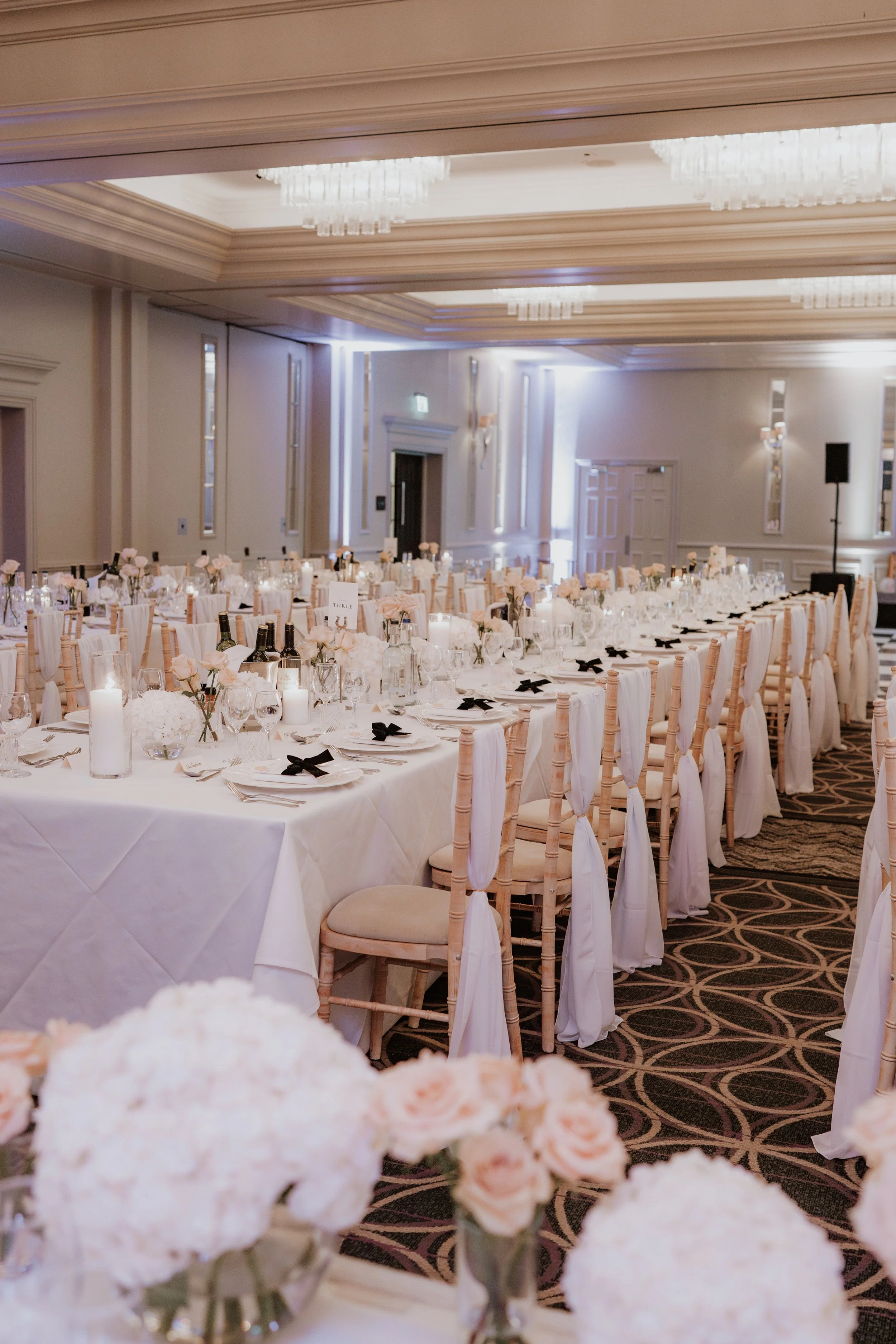 Elegant banquet hall decorated for a wedding or formal event with long tables at Sopwell House, beige chairs wrapped with white fabric, white tablecloths, floral centerpieces, candles, and black bows on napkins.