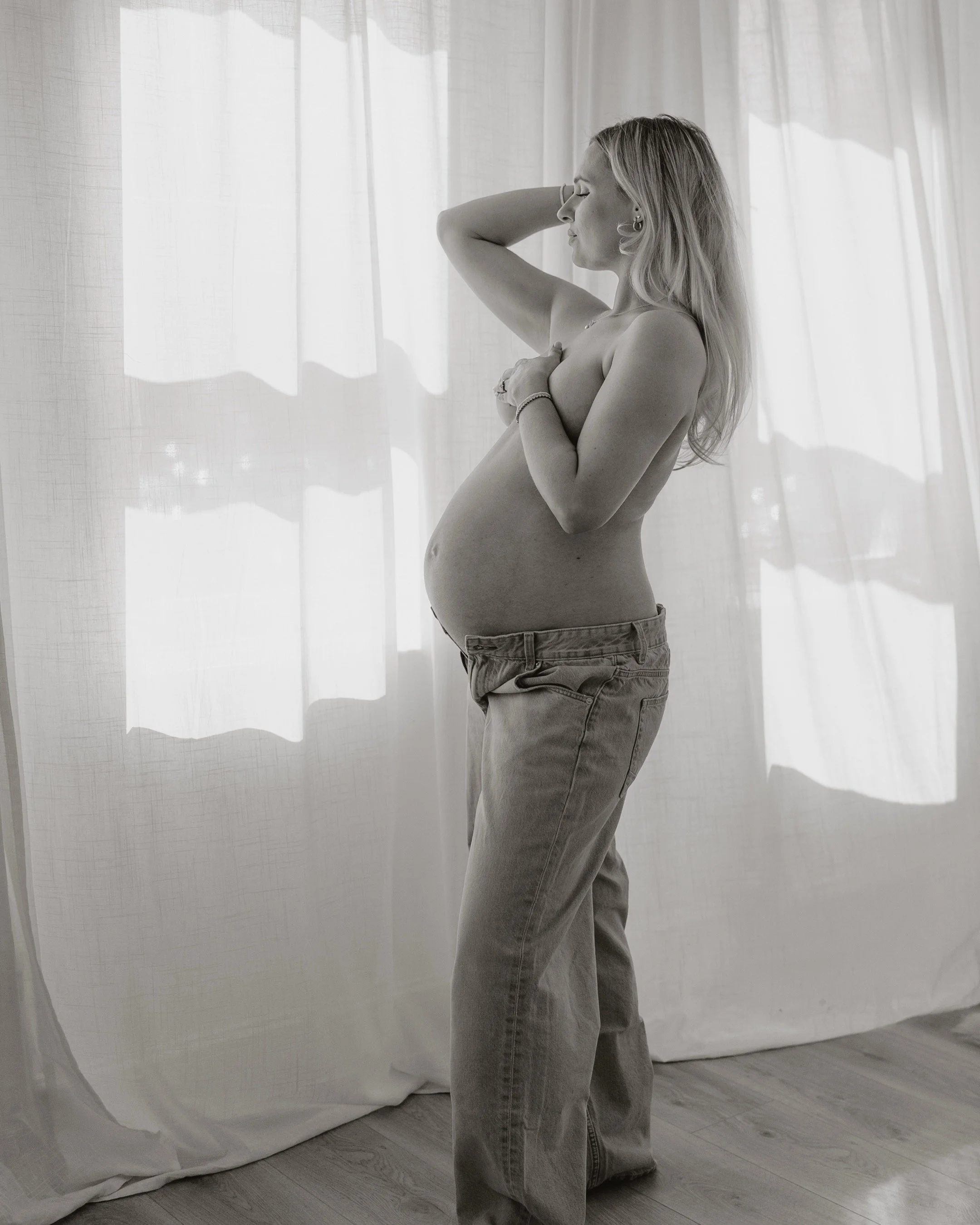 A pregnant woman standing by a window, topless, with one hand behind her head and the other crossed over her chest, wearing loose jeans.