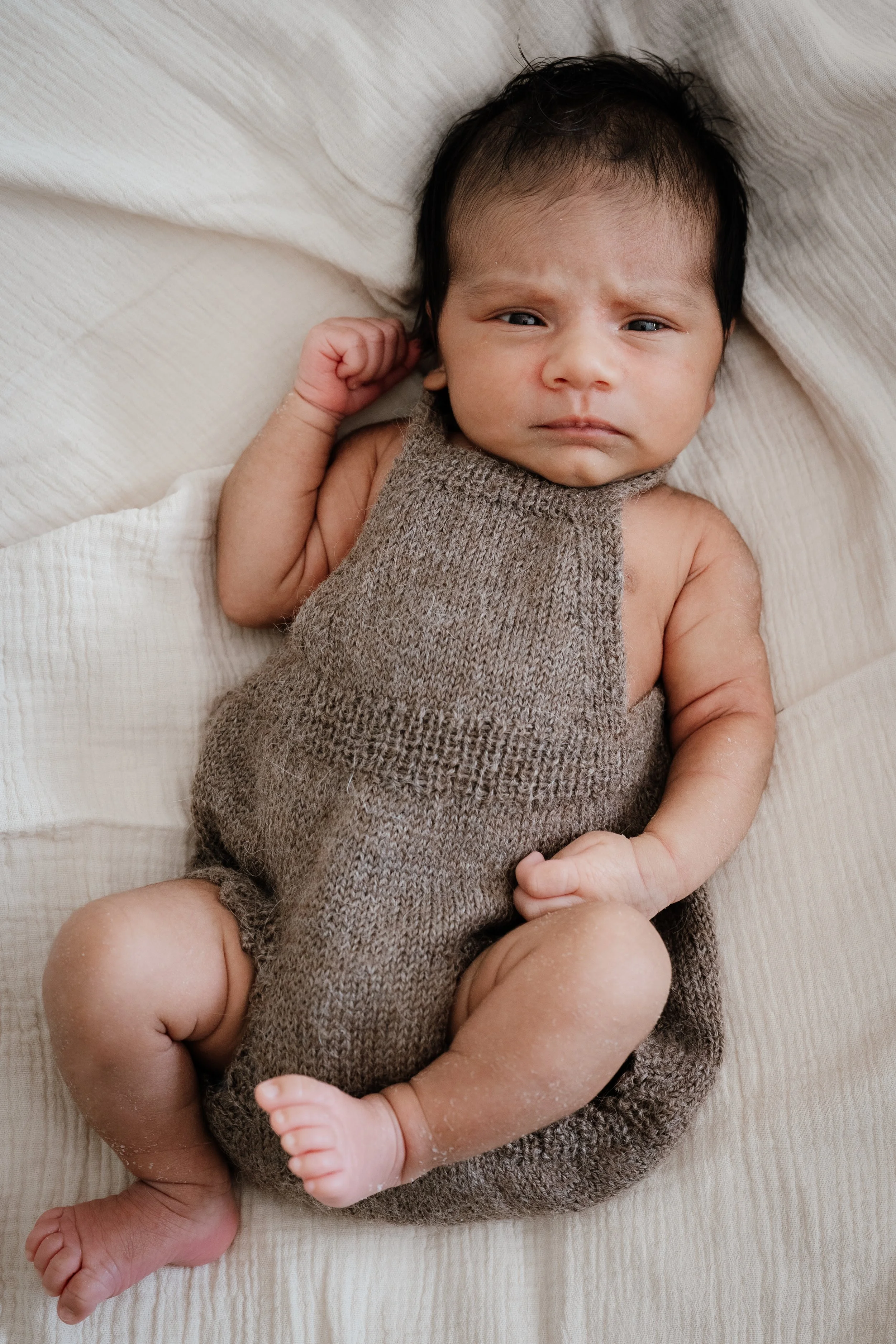 A baby lying on a cream-colored bed, wearing a knitted brown romper, with a serious expression, and short dark hair.