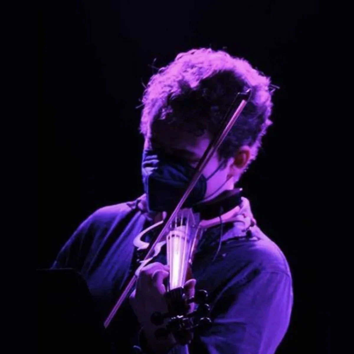 A person playing the violin on stage illuminated by purple light, wearing glasses and a face mask.